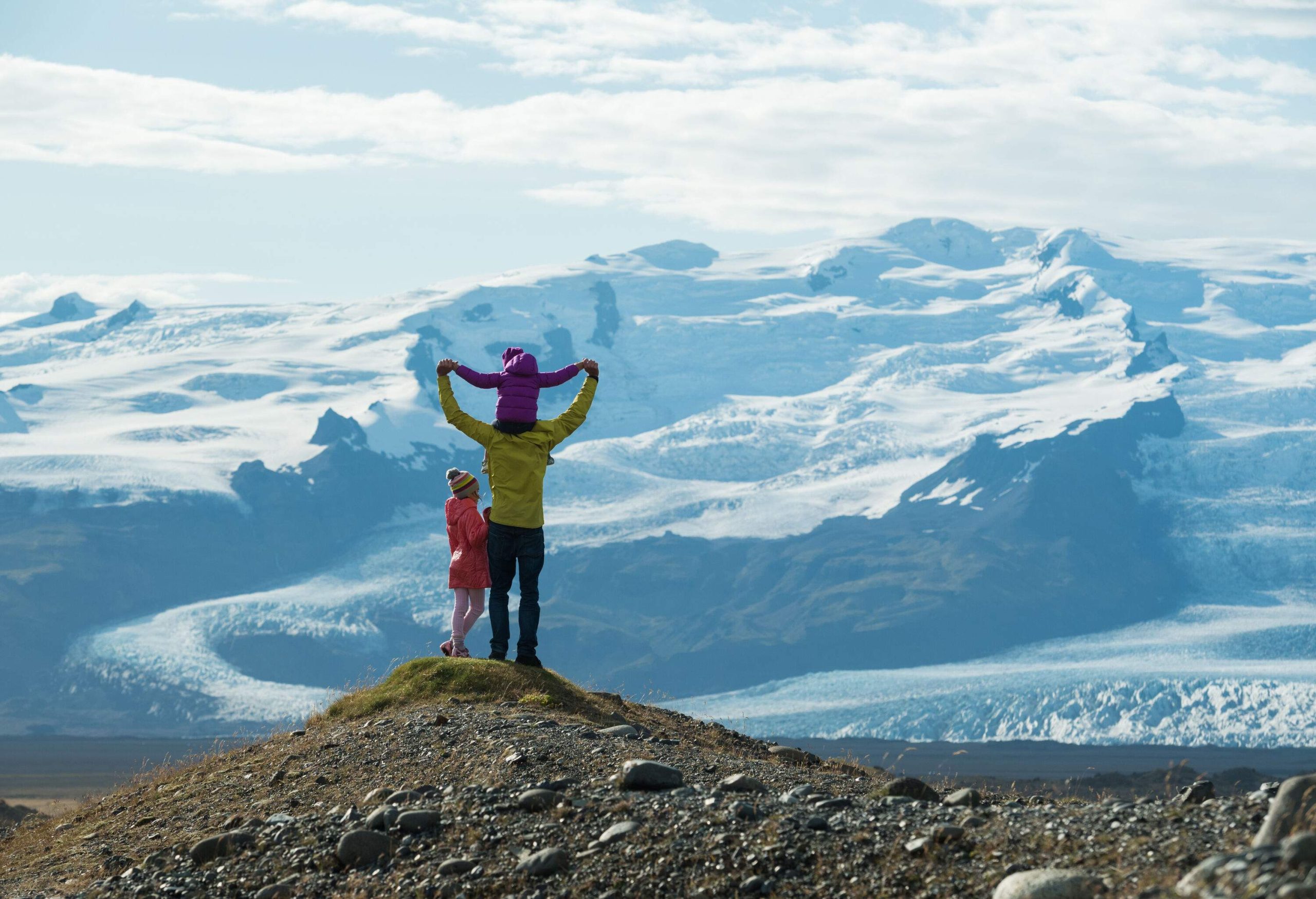 A man with a kid on his shoulders standing next to a young girl on a hilltop overlooking a glacier.