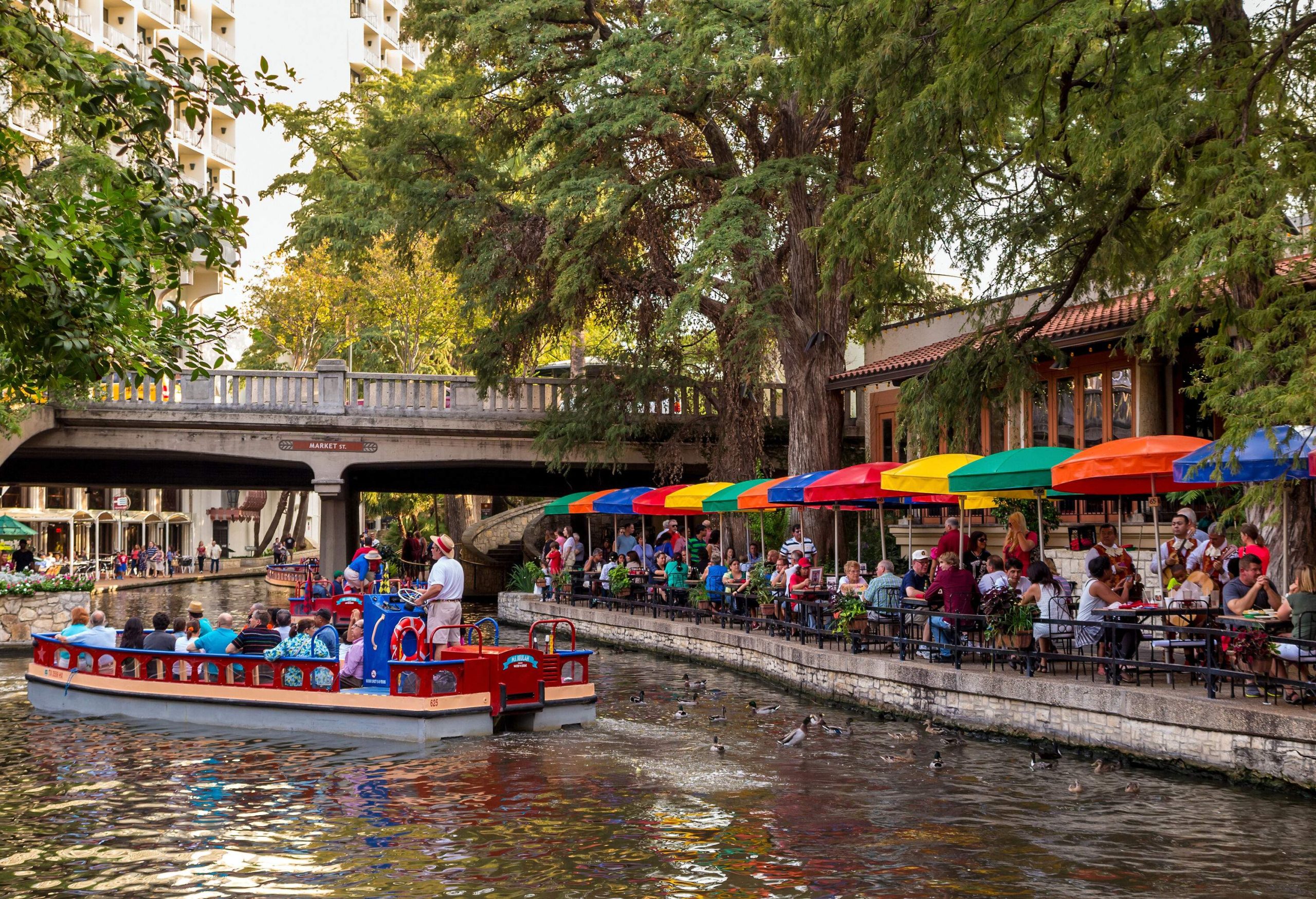 A vibrant and colourful passenger boat gracefully cruises beneath a charming bridge, while the lively riverwalk bustles with outdoor dining options, where a crowd of people enjoy the ambiance, all set against a breathtaking canopy of trees.