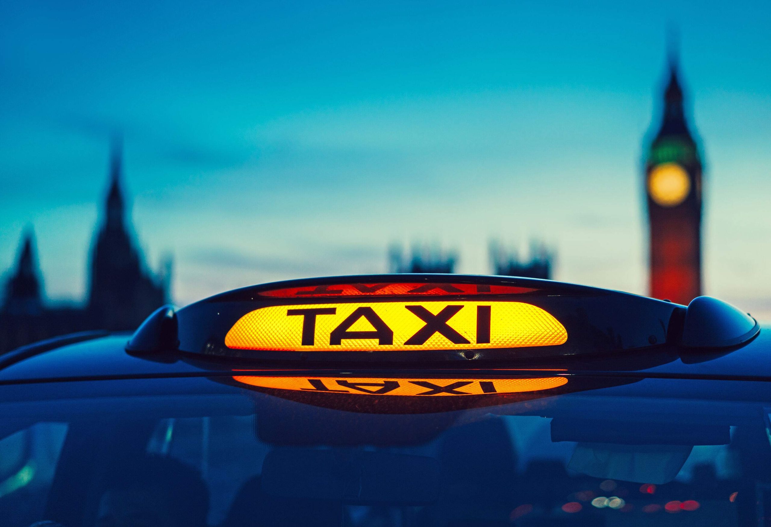 Close-up of an illuminated London taxi sign with a blurry image of Big Ben in the background.