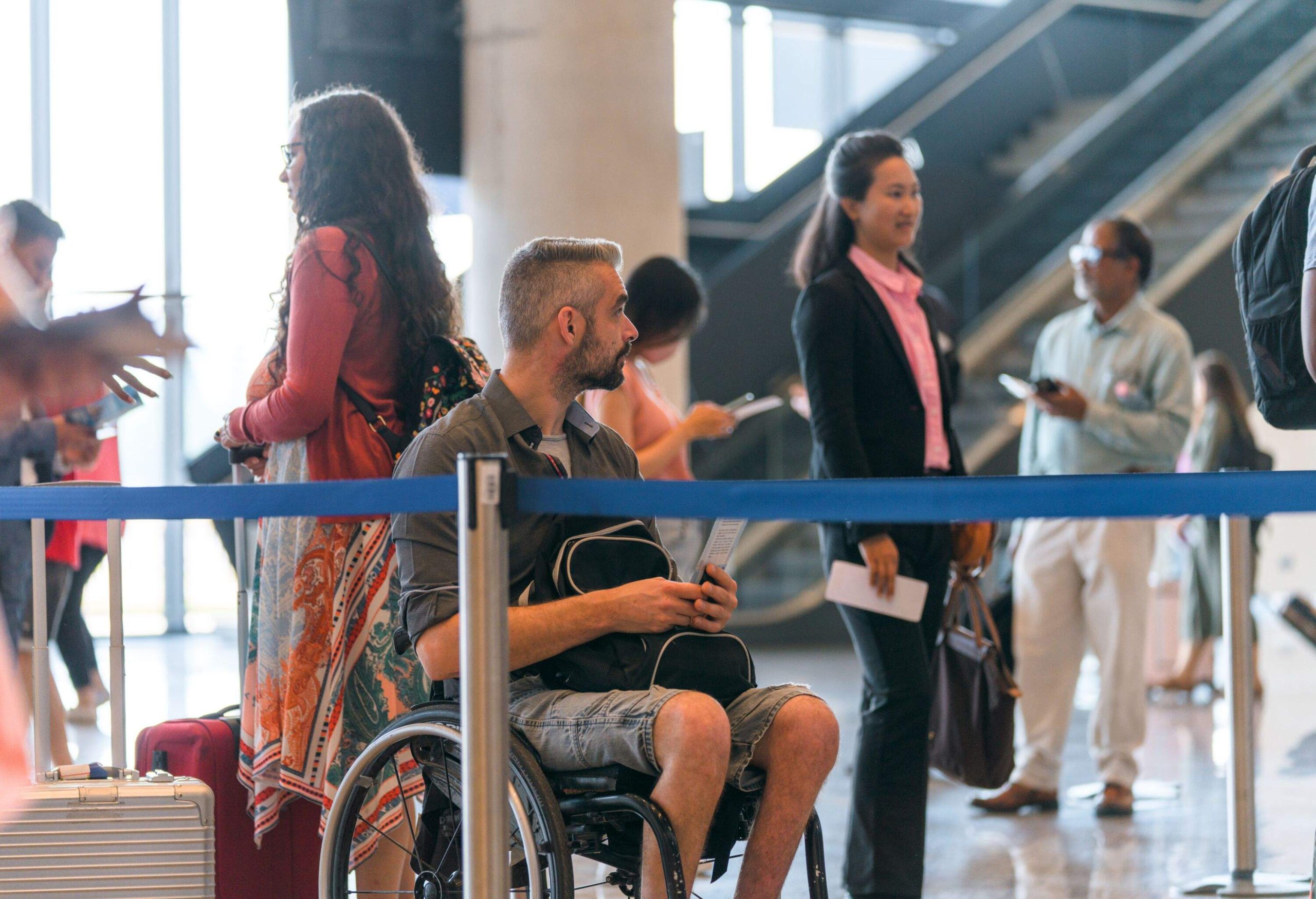 Disabled Caucasian male on a wheelchair traveling. Sitting on a wheelchair waiting in a check in line at the airport. He is wearing a protective face mask.