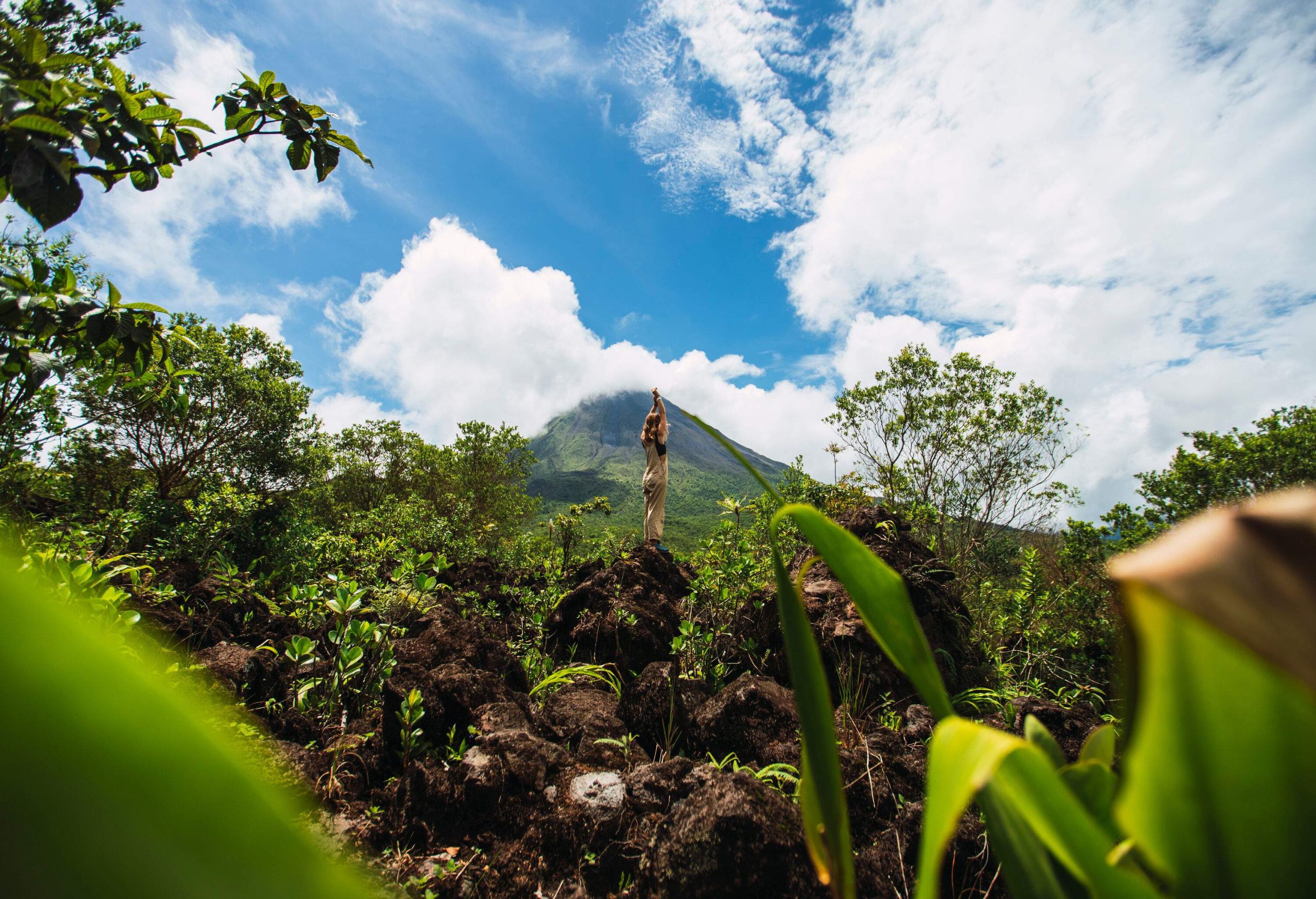 A woman standing on a rock with her hands above her head, with views of a volcano in the distance.