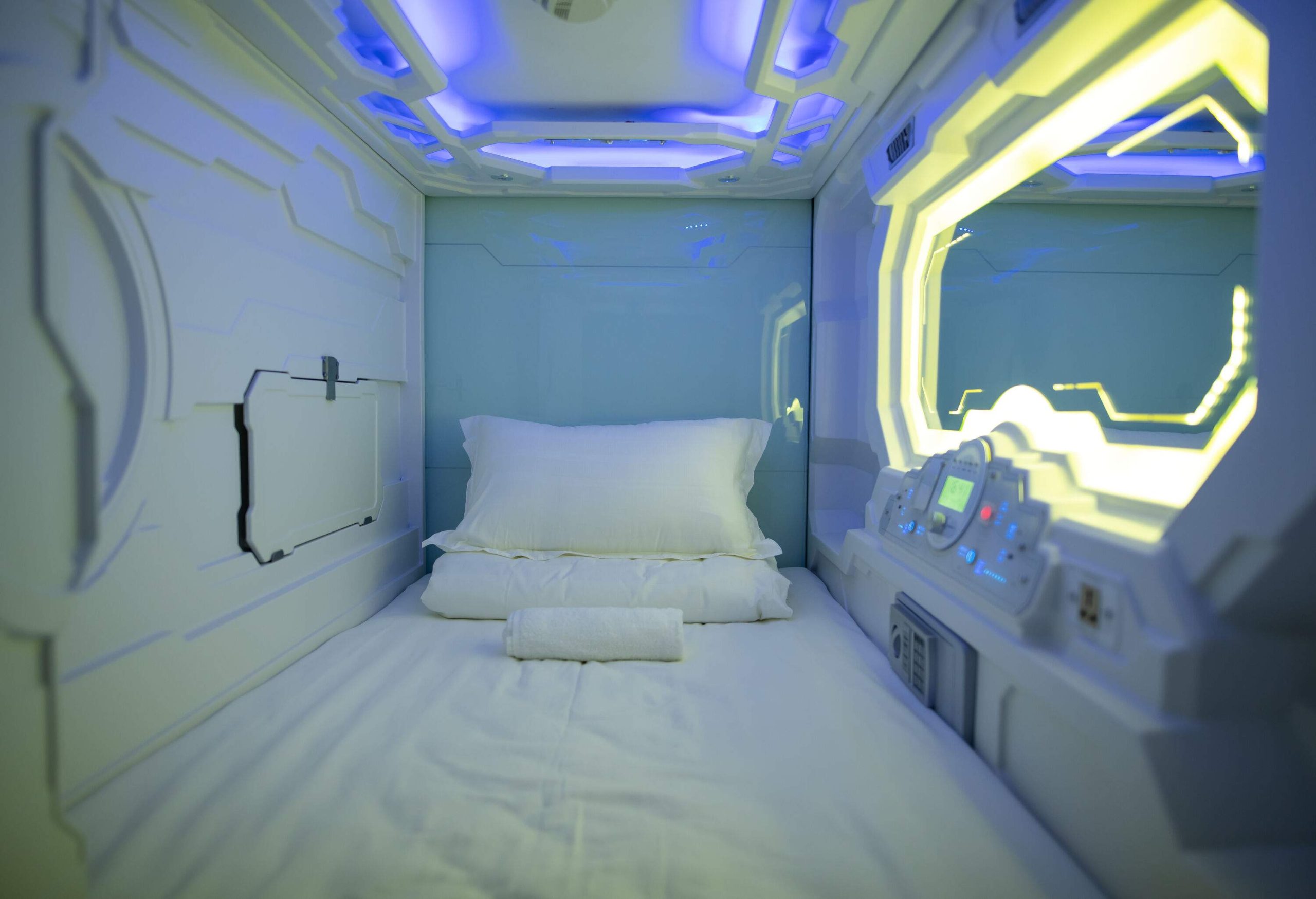 A modern and cosy capsule hotel room with white walls, equipped with an illuminated mirror and a small safe box.