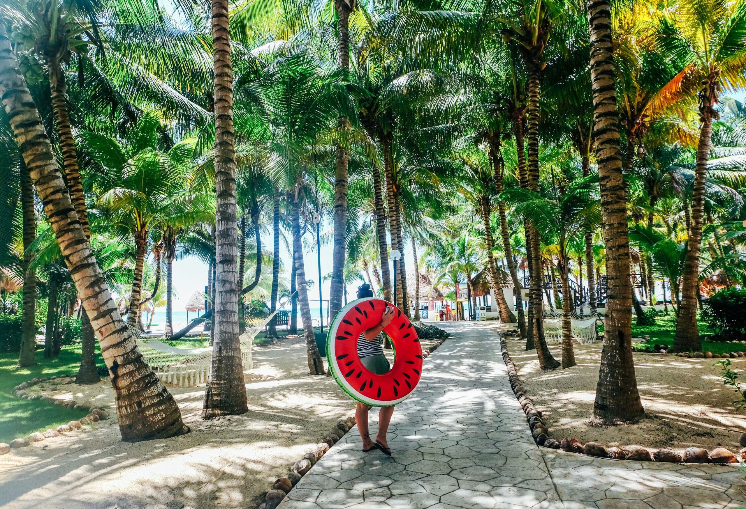 person walking through palm trees in tropical environment holding a large red inflatable ring