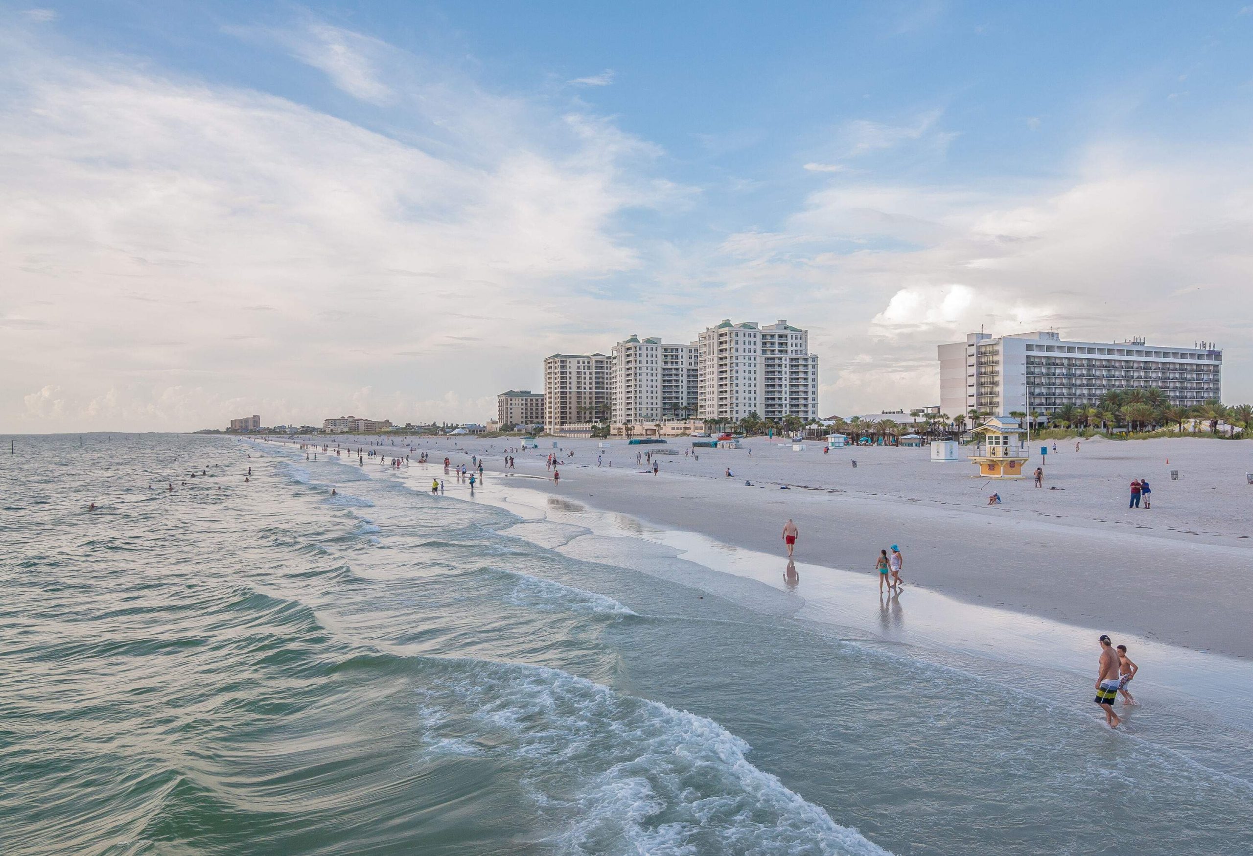 A beach with rhythmic waves, beachgoers leisurely walking by the water's edge, and distant buildings adding urban allure to the scenic panorama.