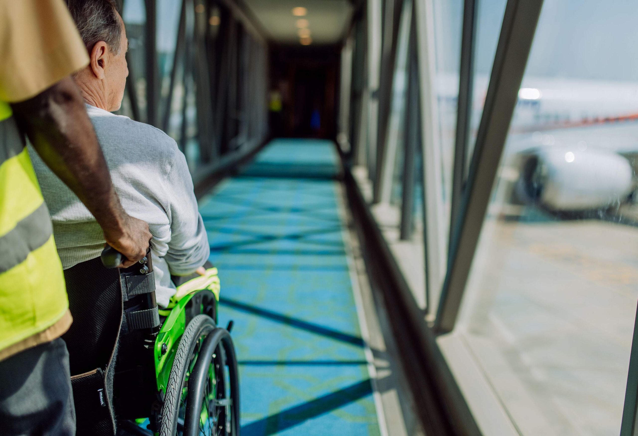 An airport worker pushes a man in a wheelchair inside an airport passenger boarding bridge.