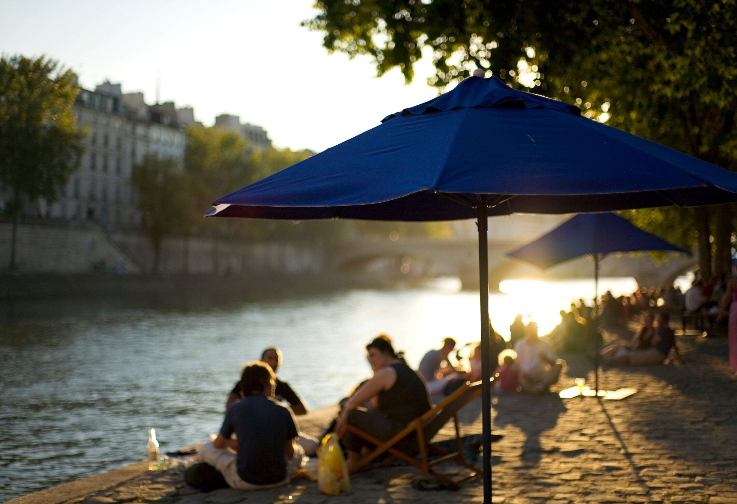 Bokeh of people sitting under blue umbrellas by the river bank.