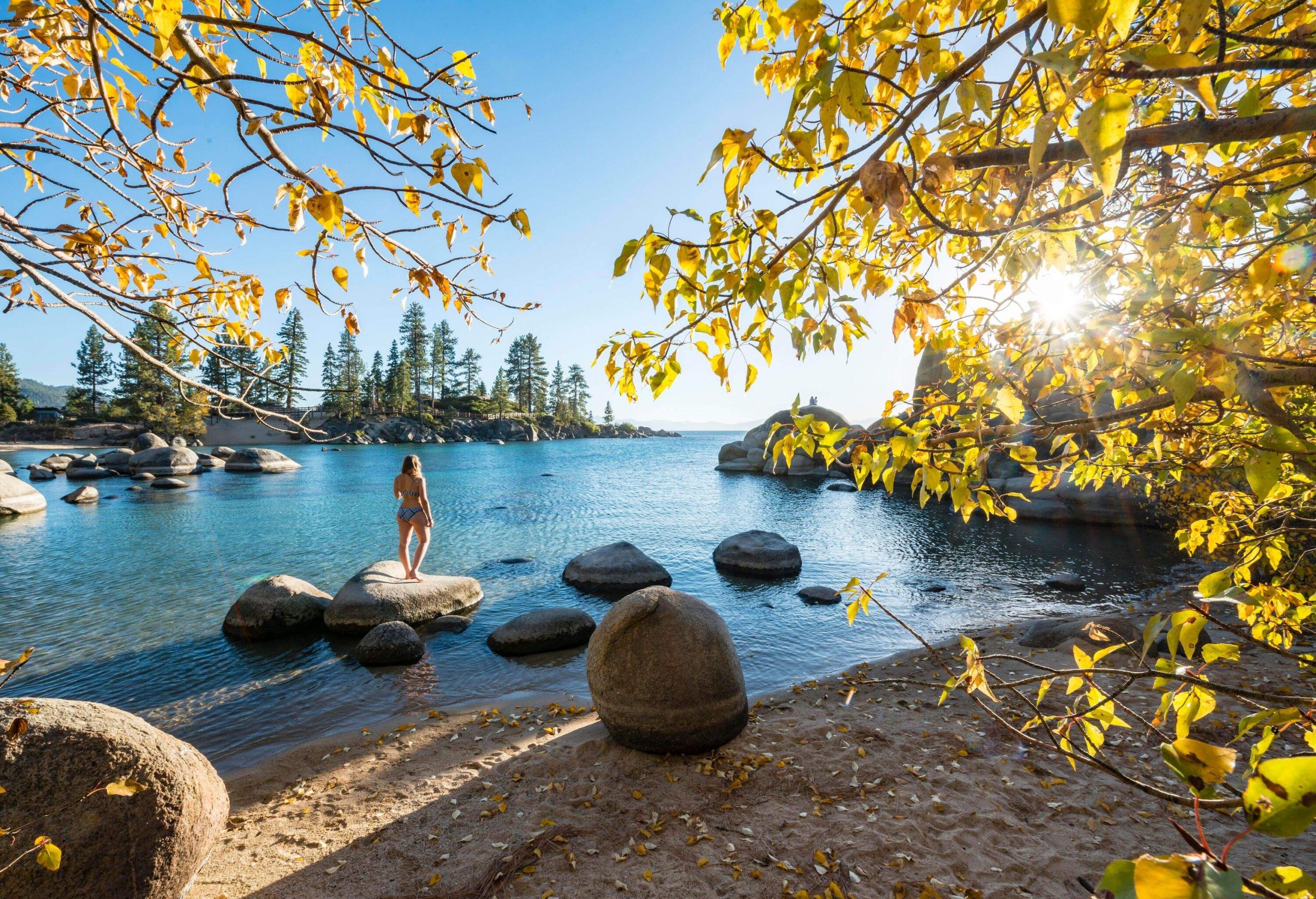 A person standing on the boulders in the shallow part of the lake, with the sun shining through the yellow autumn leaves in the foreground.
