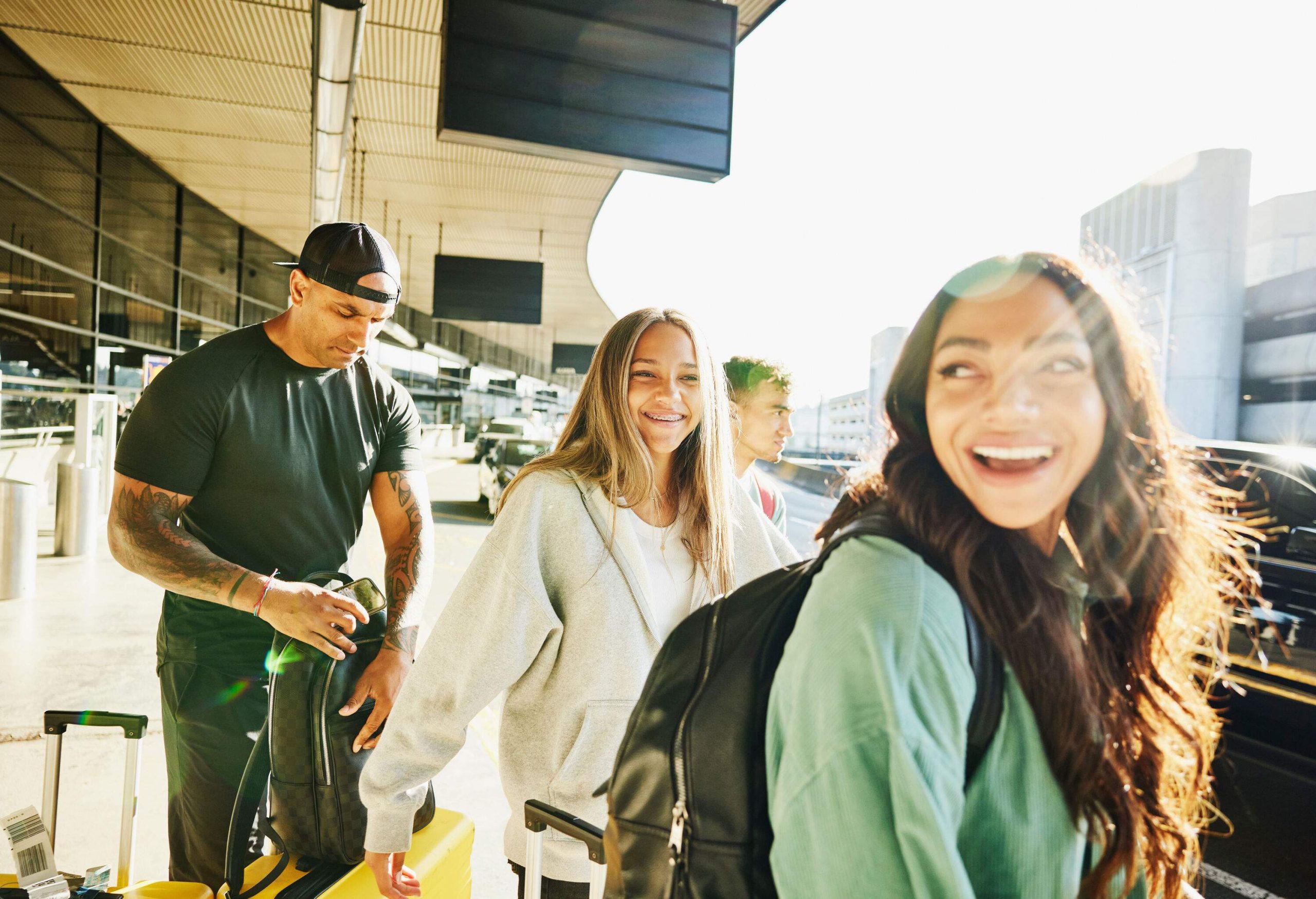 A group of happy people with their luggage stands on the curbside of an airport.