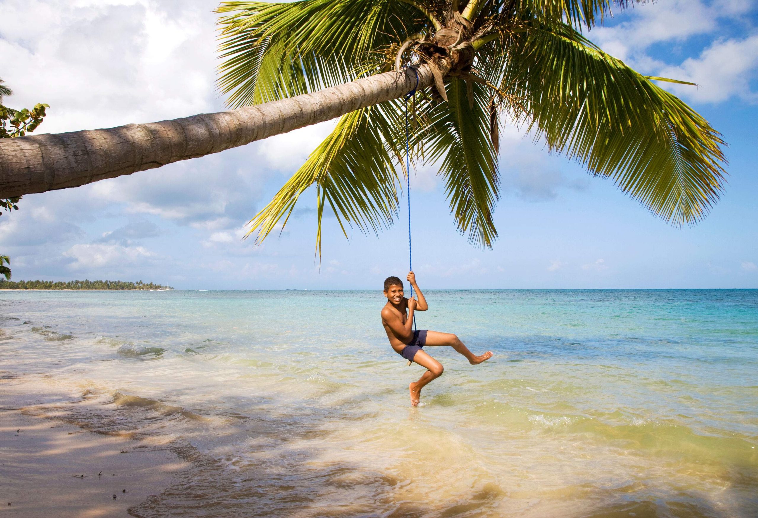 Boy hanging over a palm tree by a Caribbean beach smiling at the camera