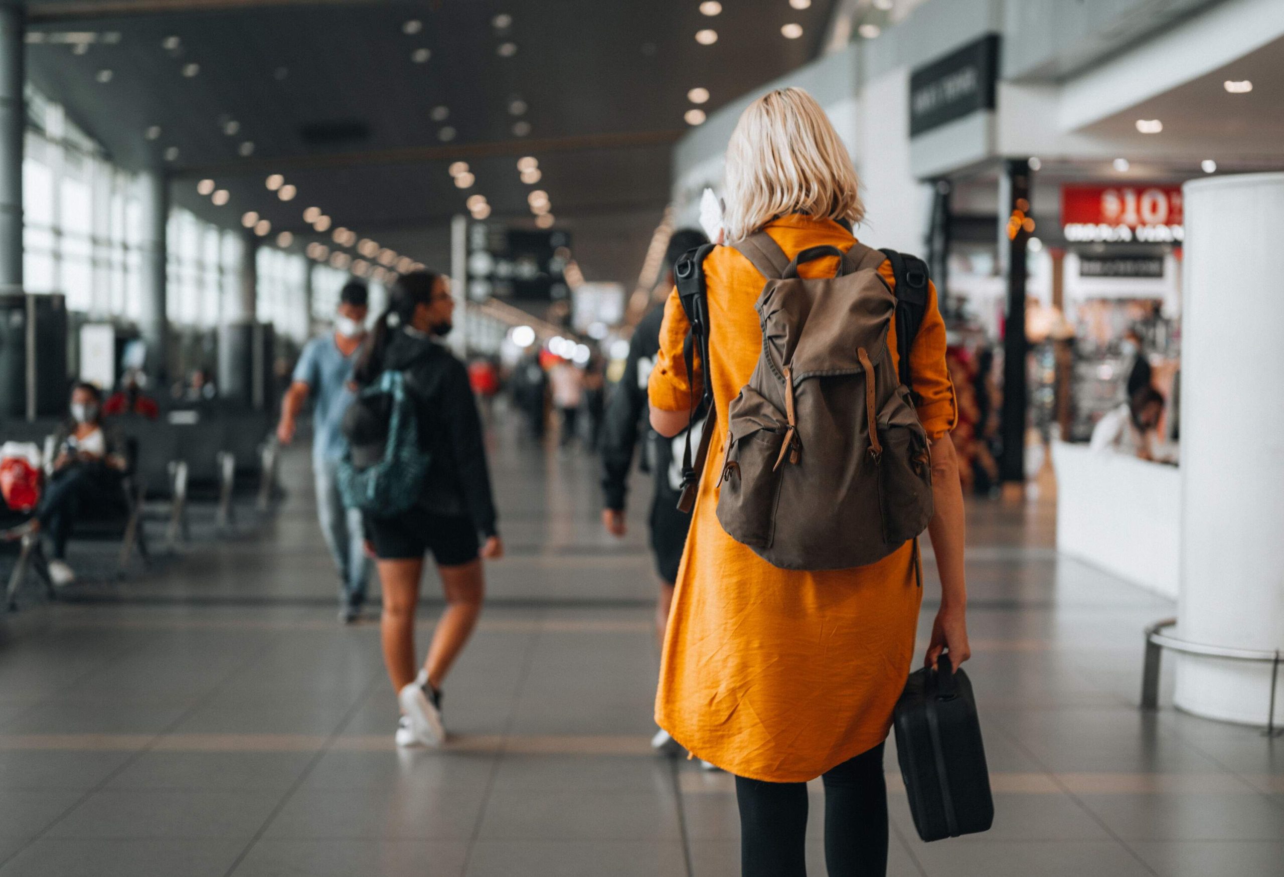 A lady strolling through the airport terminal carrying several bags, surrounded by shops and cafés.