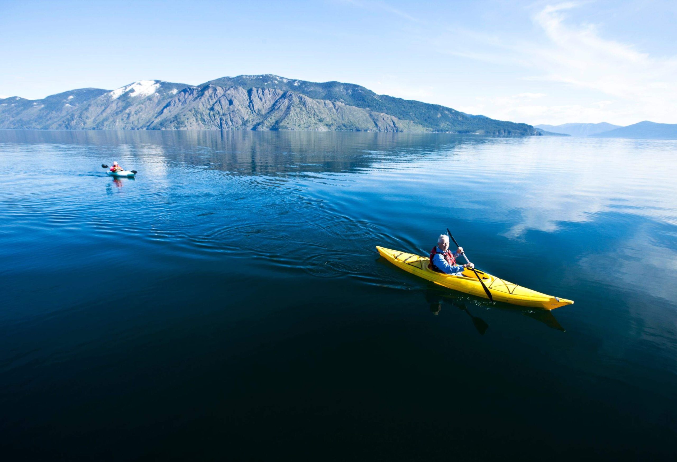 A couple in different yellow kayaks on a lake by a mountain on a sunny day