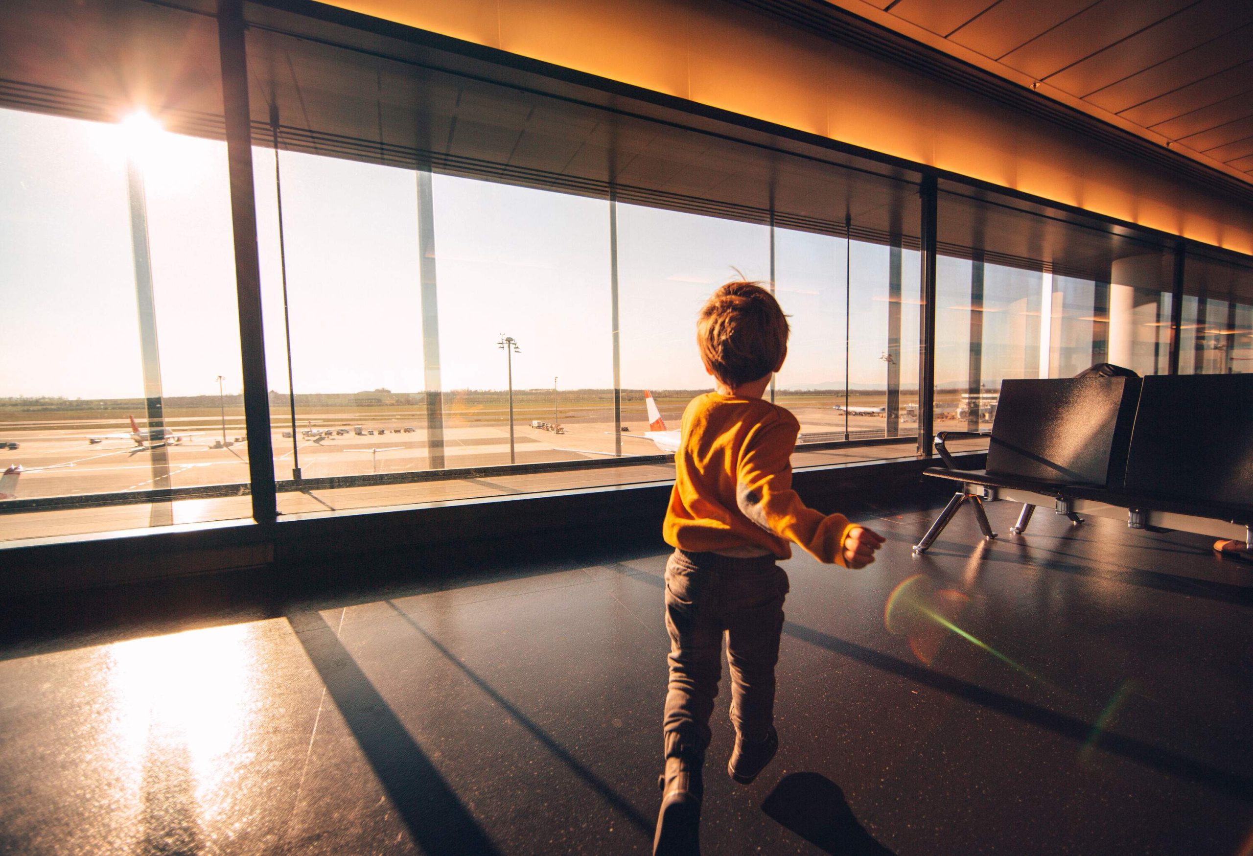 A young child in orange sweater runs on an airport lounge with large windows.
