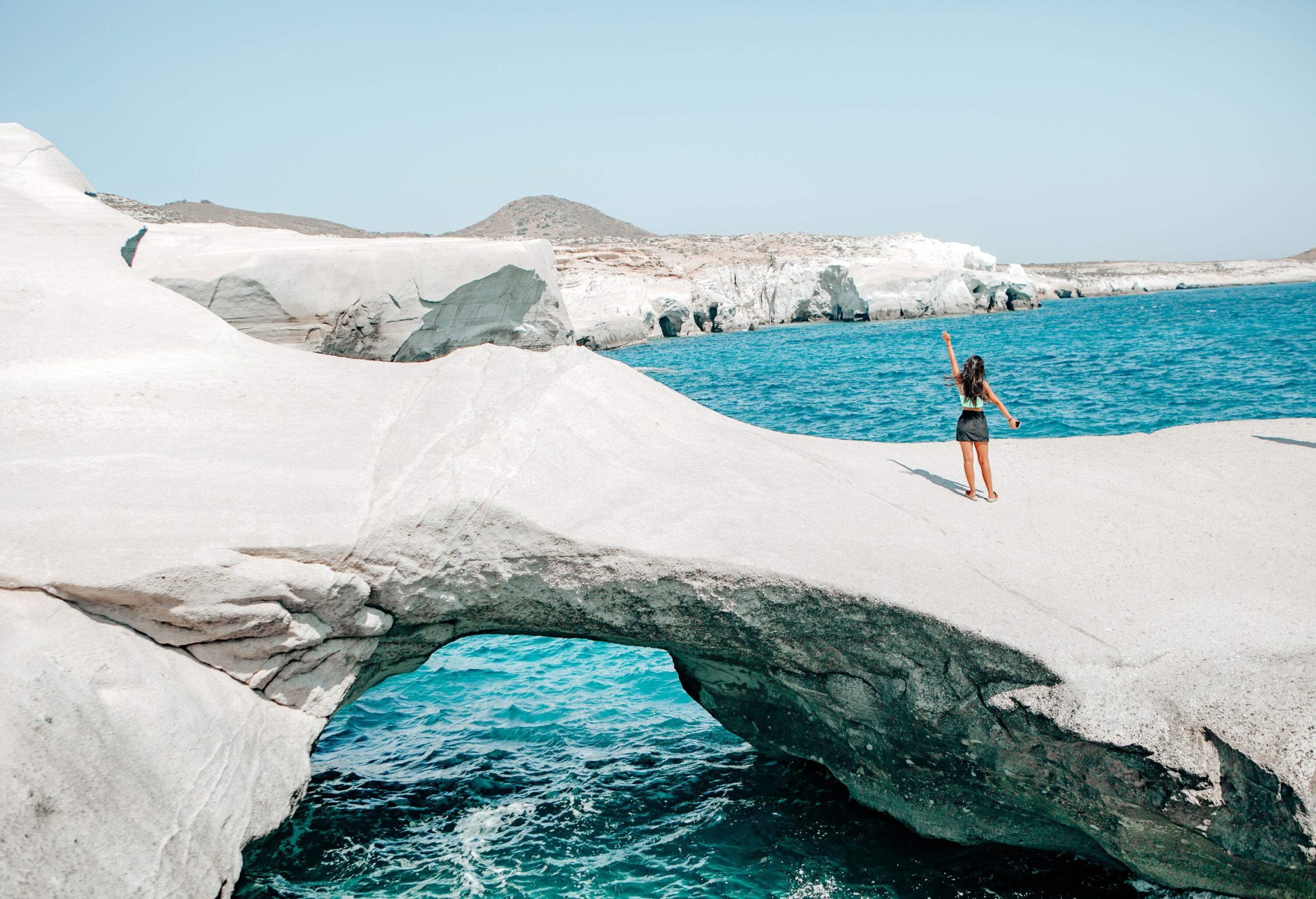 A woman standing on a natural arch formation that plunges into the sea.