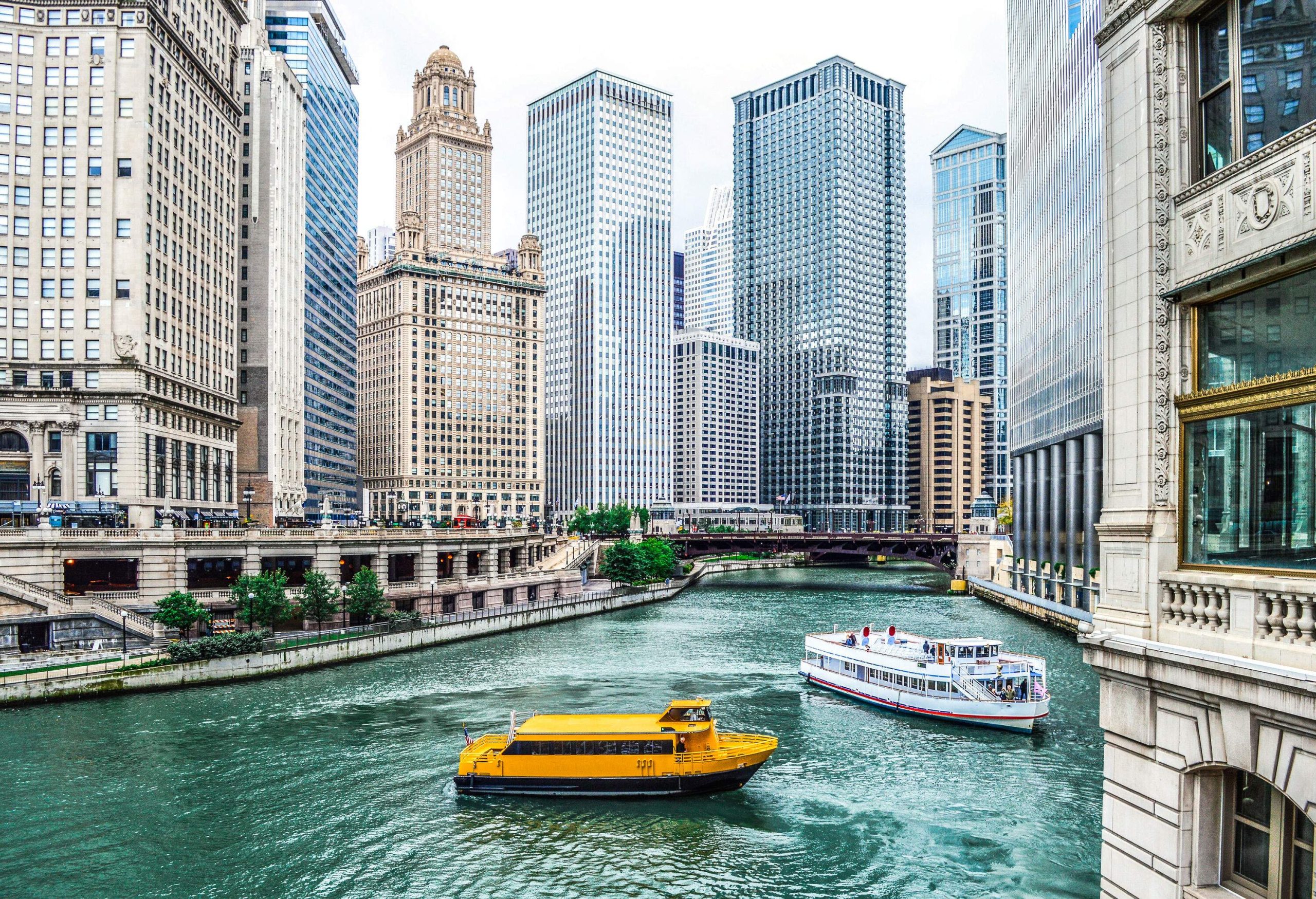 Two passenger boats are travelling the canal in the middle of a cityscape filled with high-rise structures.