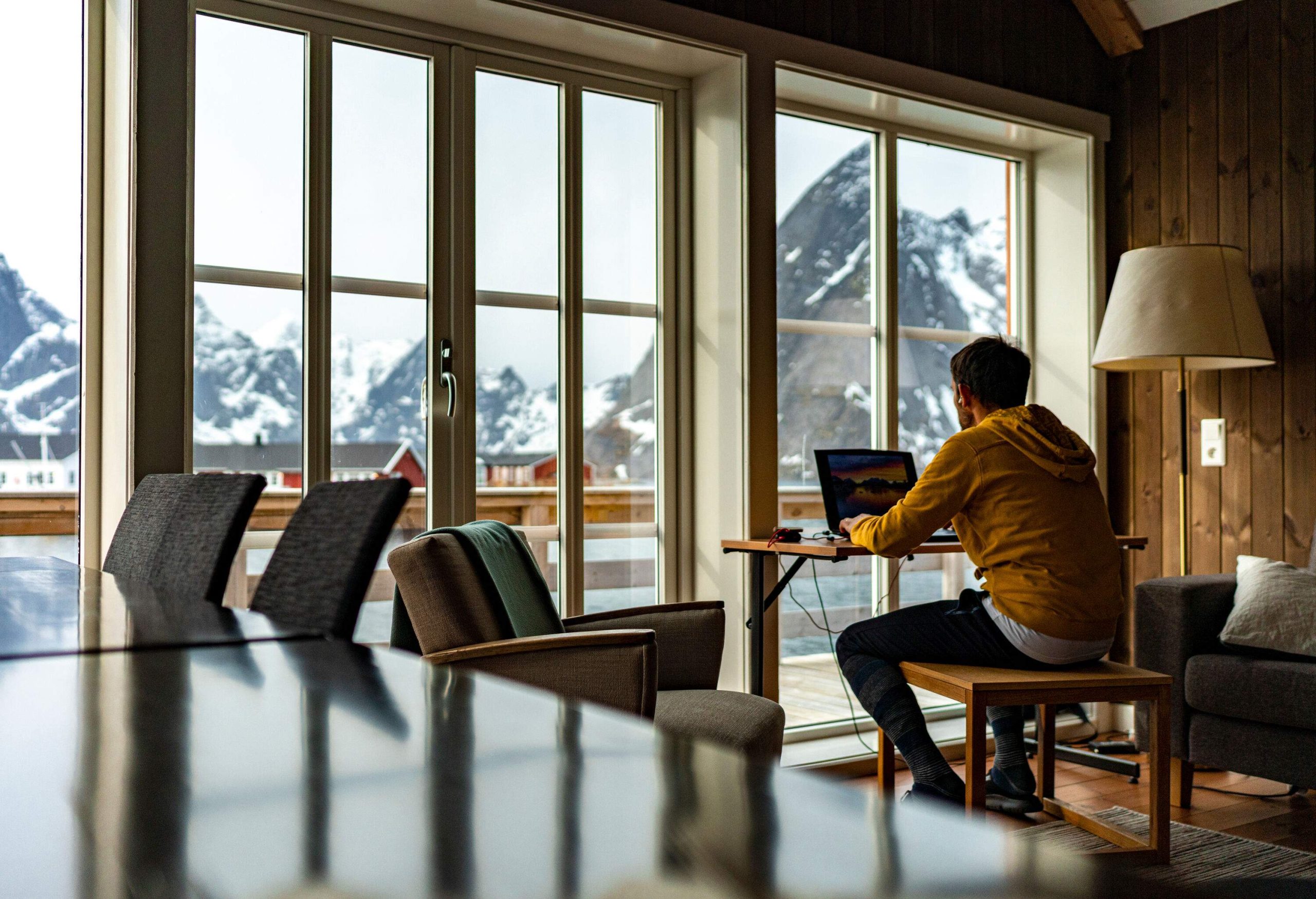 A man wearing a yellow sweatshirt working on his computer with incredible views of the snowy mountains outside.