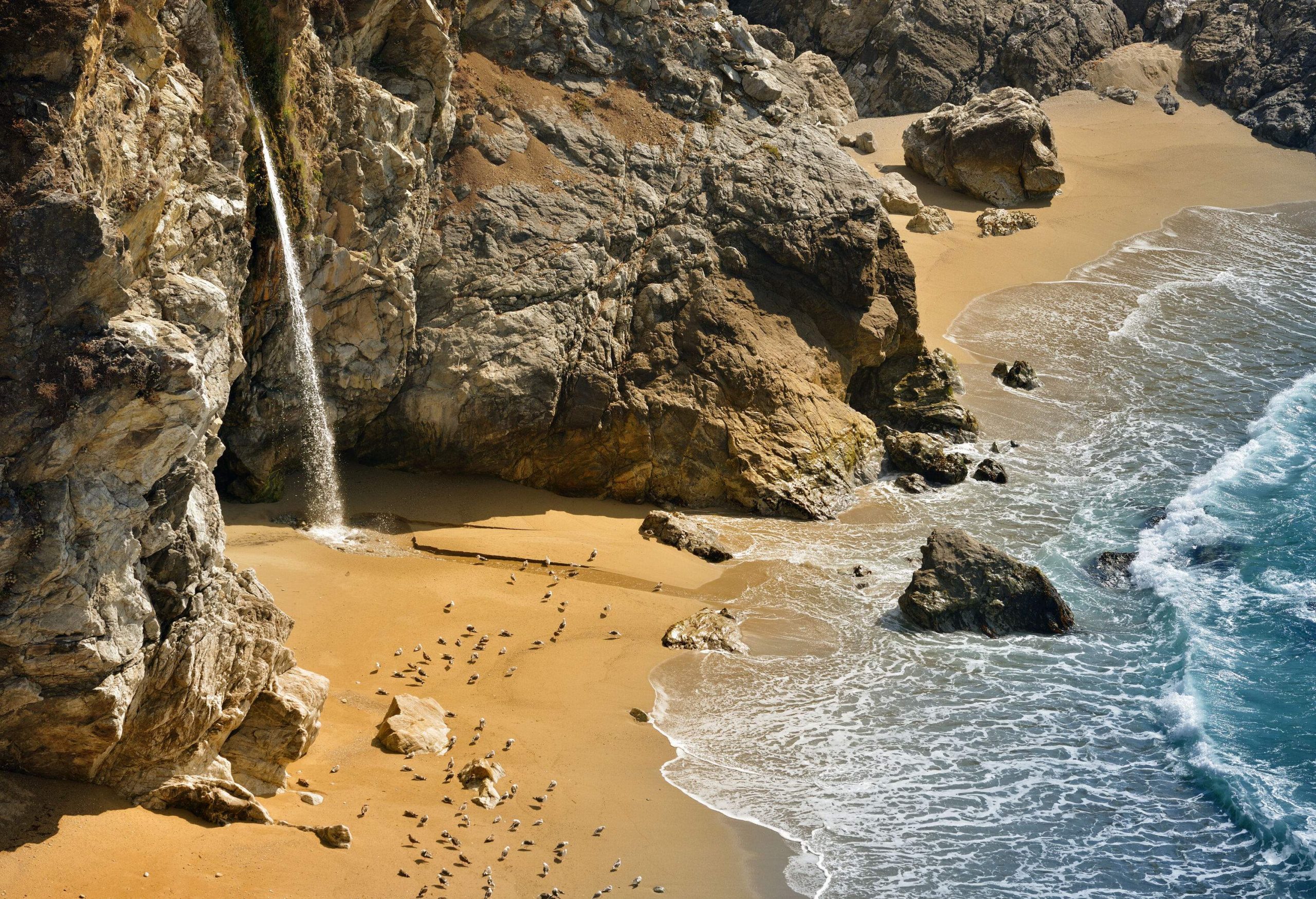 A rocky bay with a flock of seabirds on its sandy beach and a waterfall flowing to where the waves break on the shore.