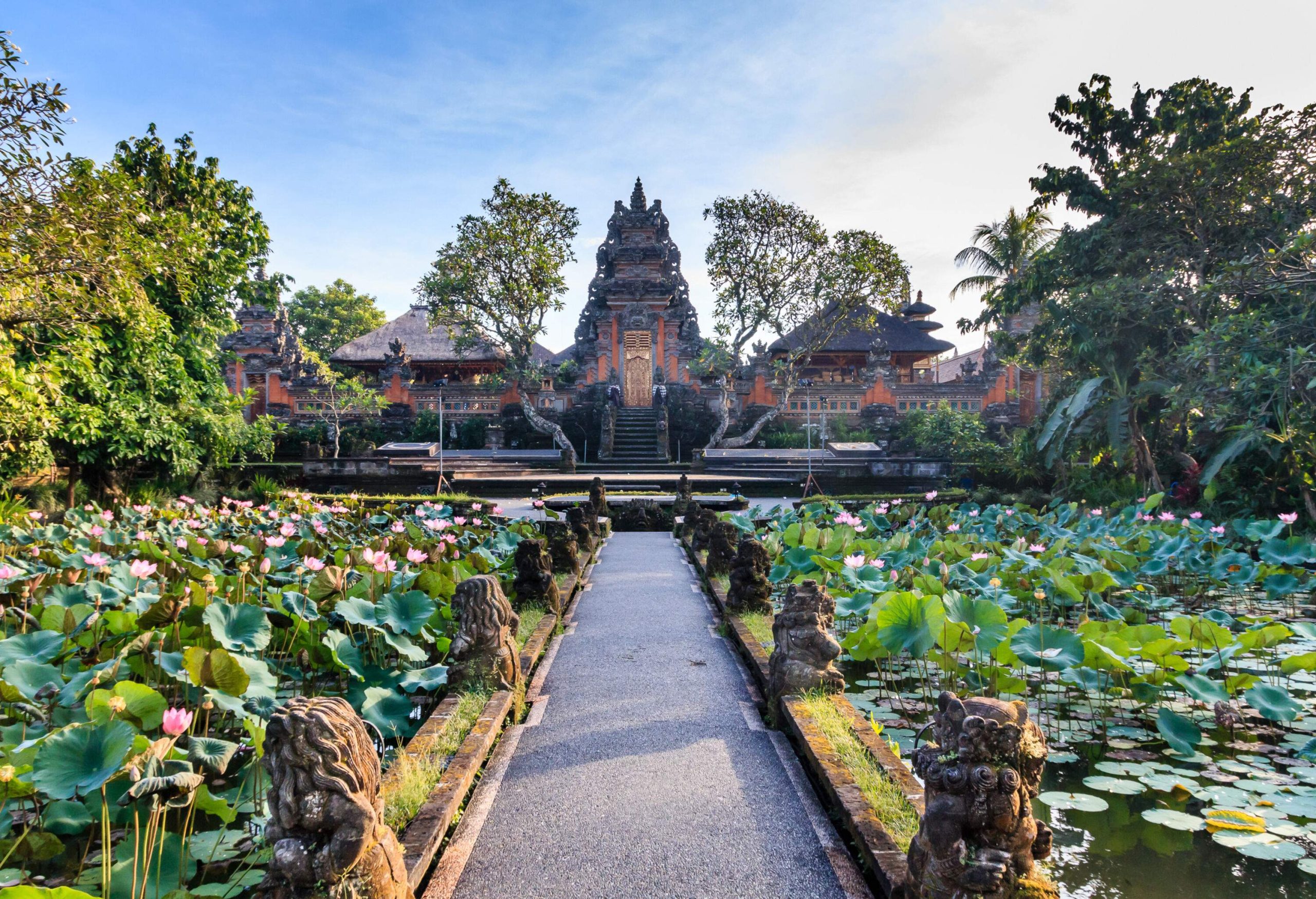 A lotus garden surrounds a walkway lined with mythical creatures that leads to the stairs of a tall gateway with golden doors situated between two cottages.