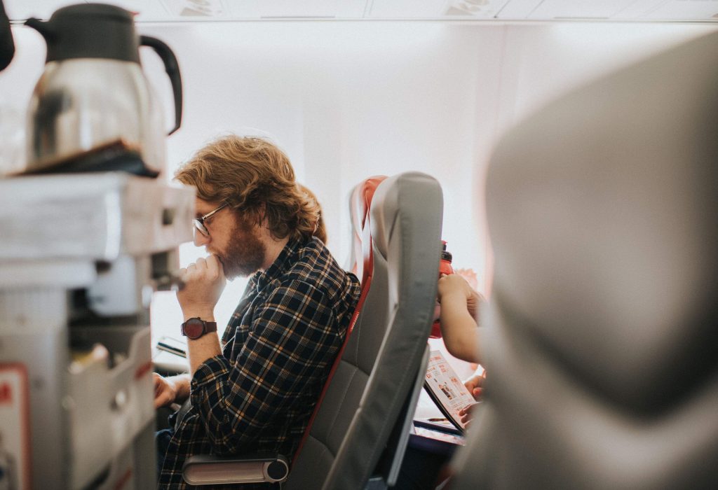 A man on a plane sits comfortably, wearing checkered long sleeves and eyeglasses, engrossed in a book.