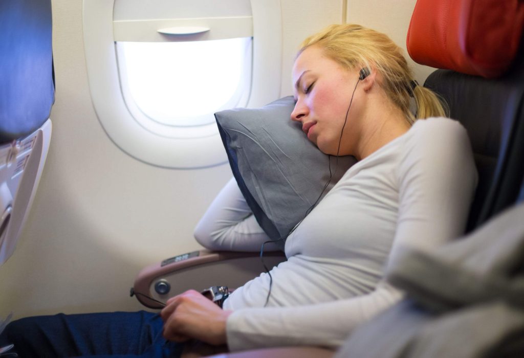 A blonde woman listens to music as she takes a nap in an airplane seat.