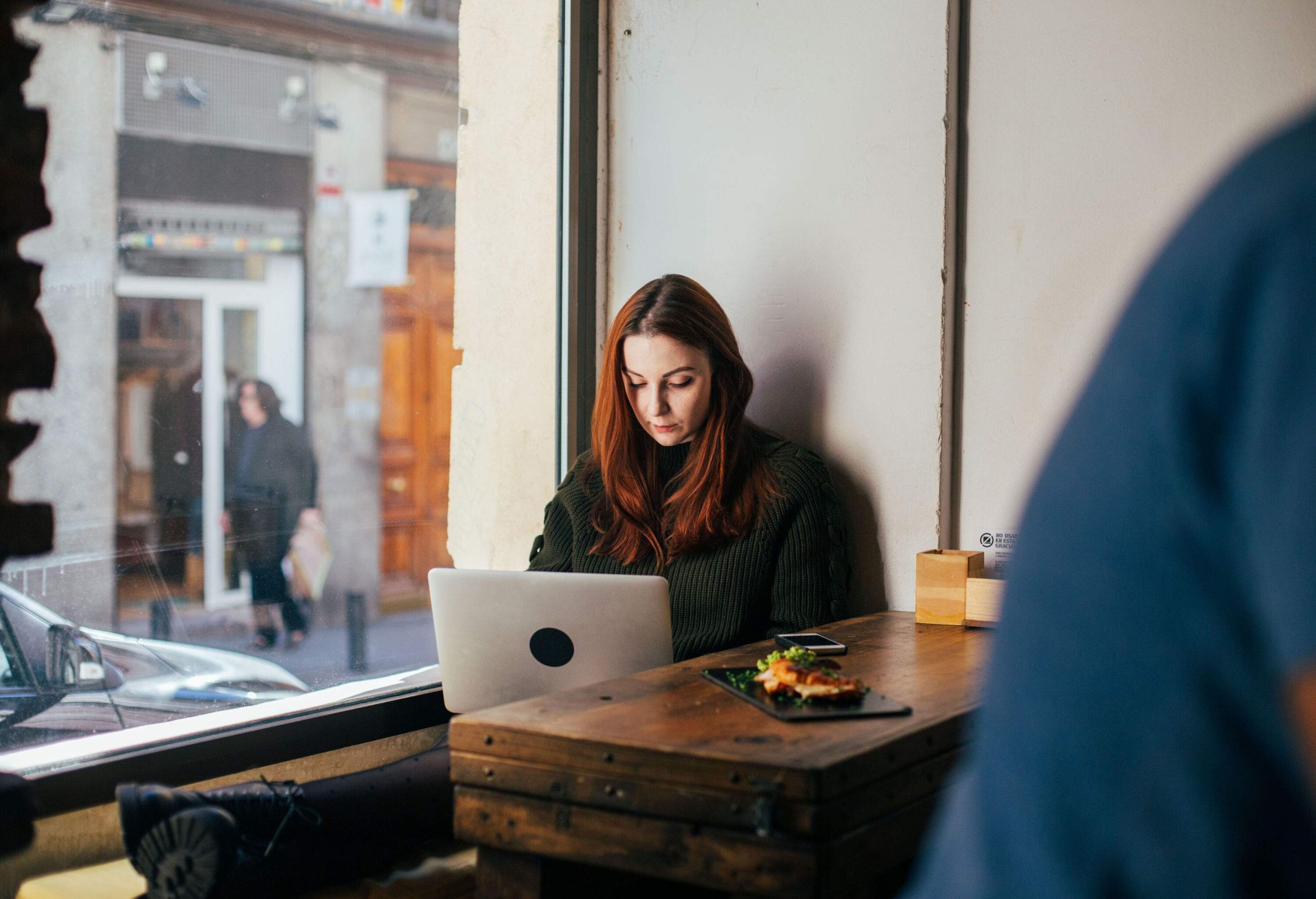 A long-haired lady sits comfortably near the big glass window working on her laptop in a cosy cafe.