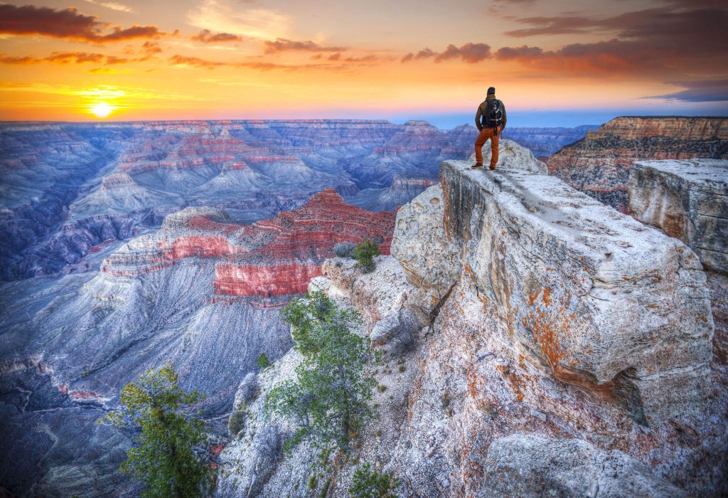 A man stands on top of a rocky mountain overlooking the Grand Canyon landscape.