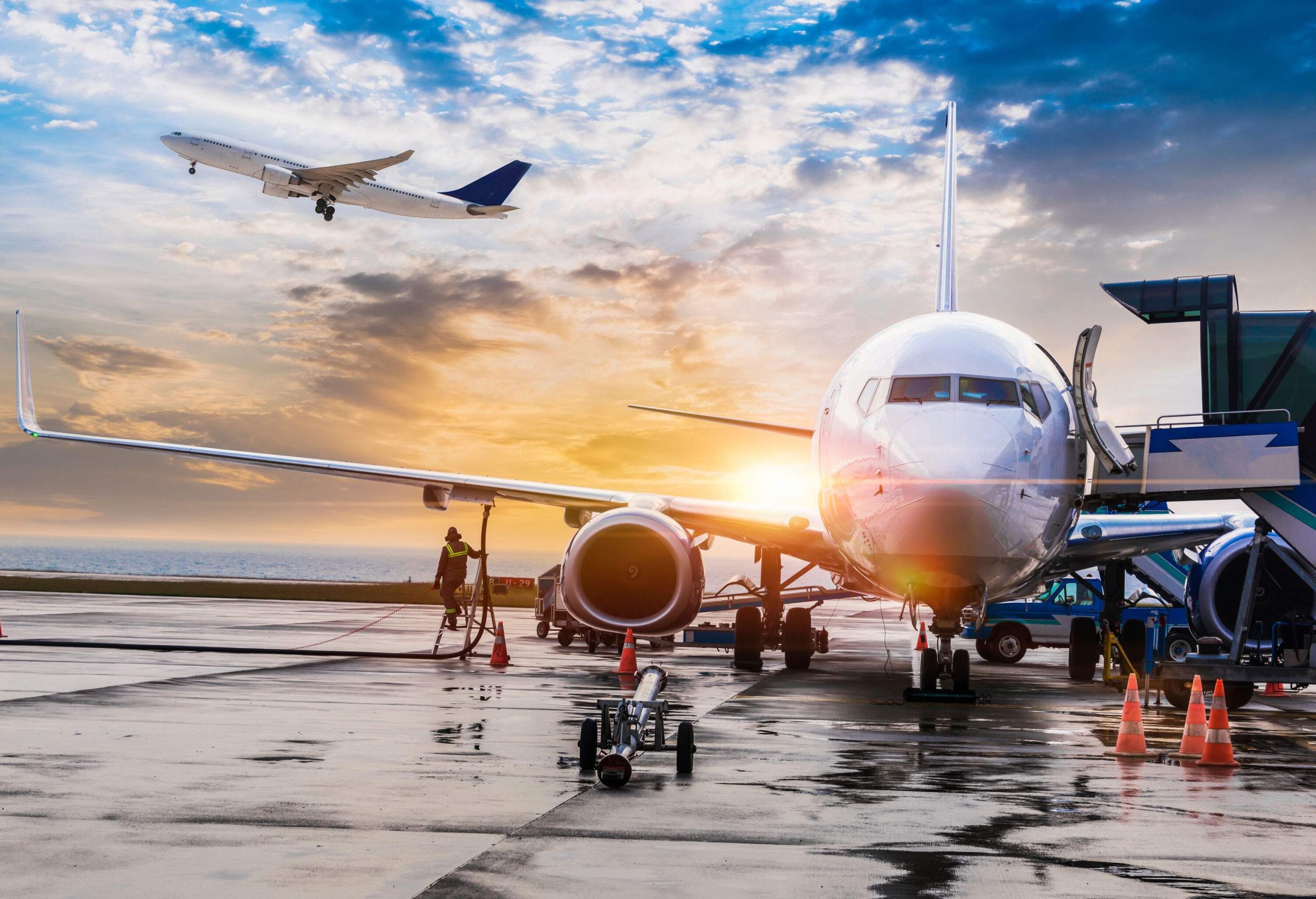 A white airplane parked on a runway with views of another airplane taking off at sunset.