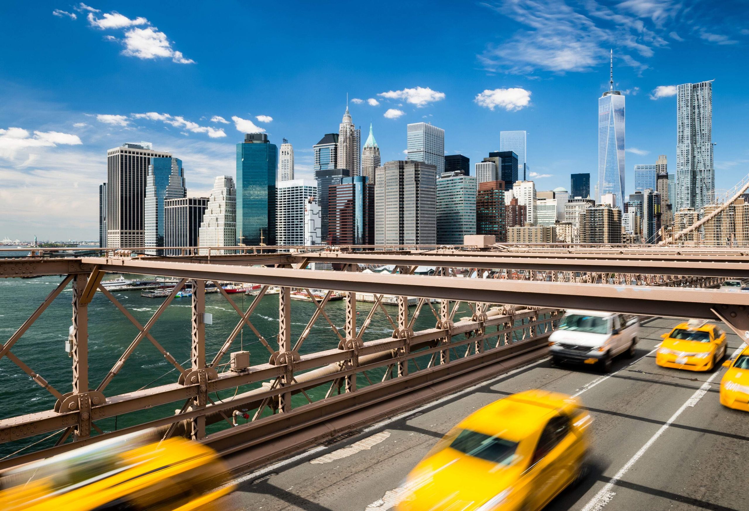 Iconic yellow taxis are in motion on a bridge overlooking the modern coastal city with towering skyscrapers.