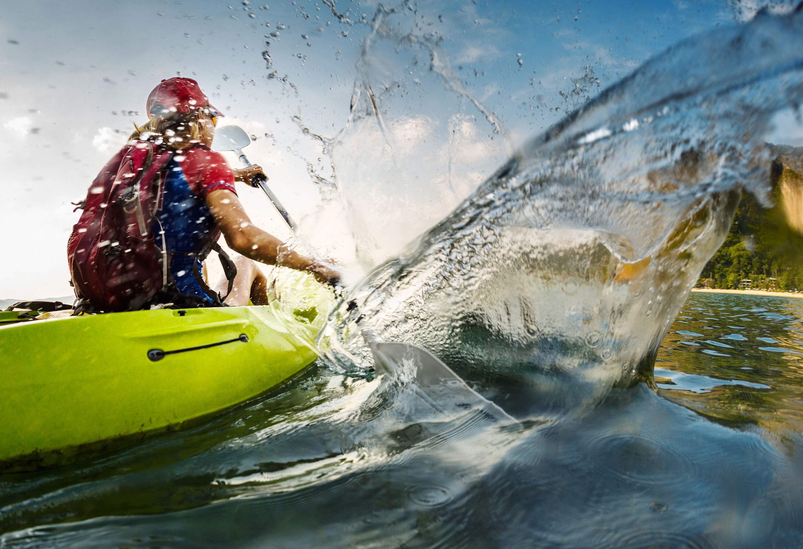 A blonde lady wearing a red cap on a kayak paddles against the water with splashes.