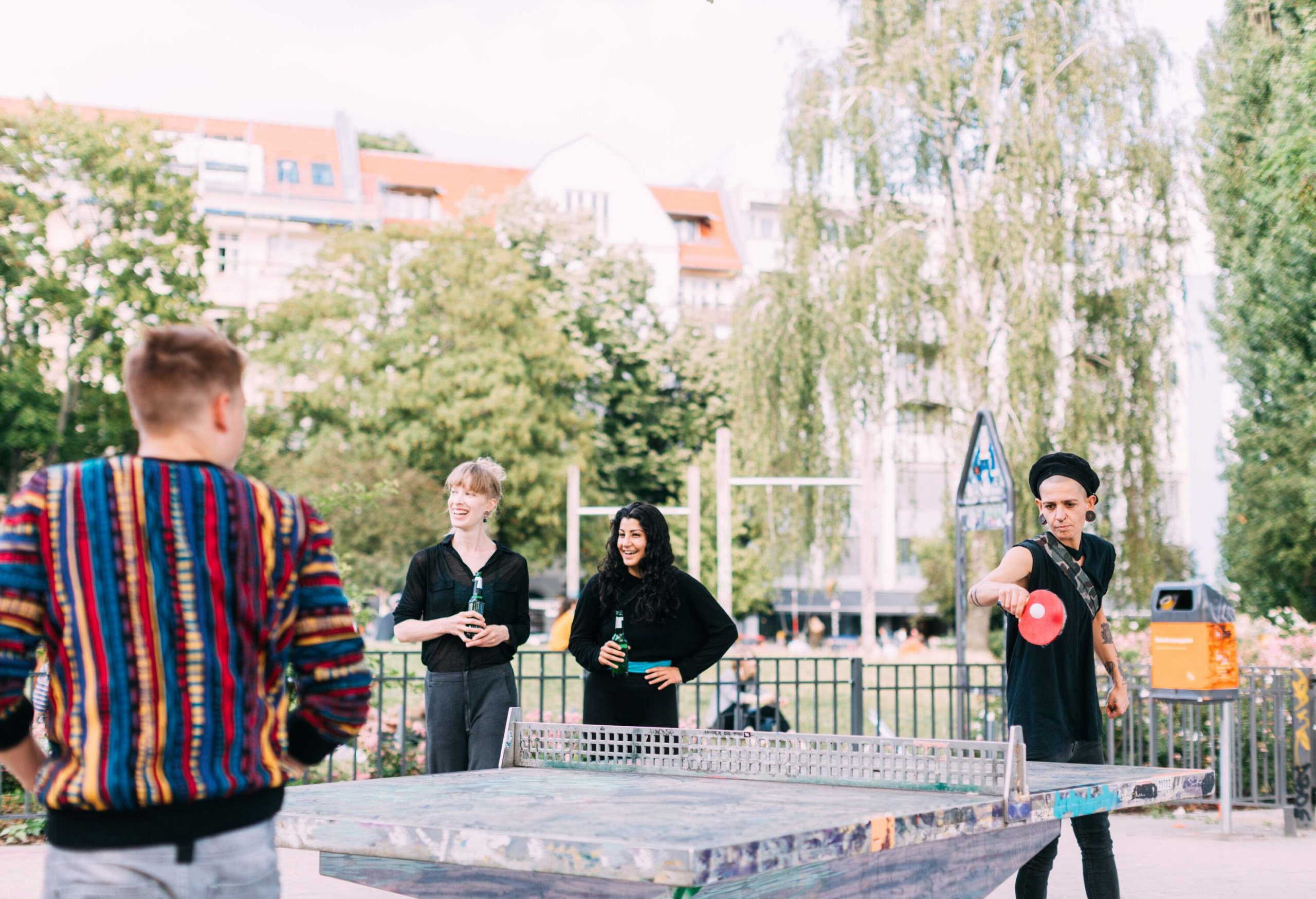 Two women with a drink in hand standing next to a couple of guys playing ping-pong.