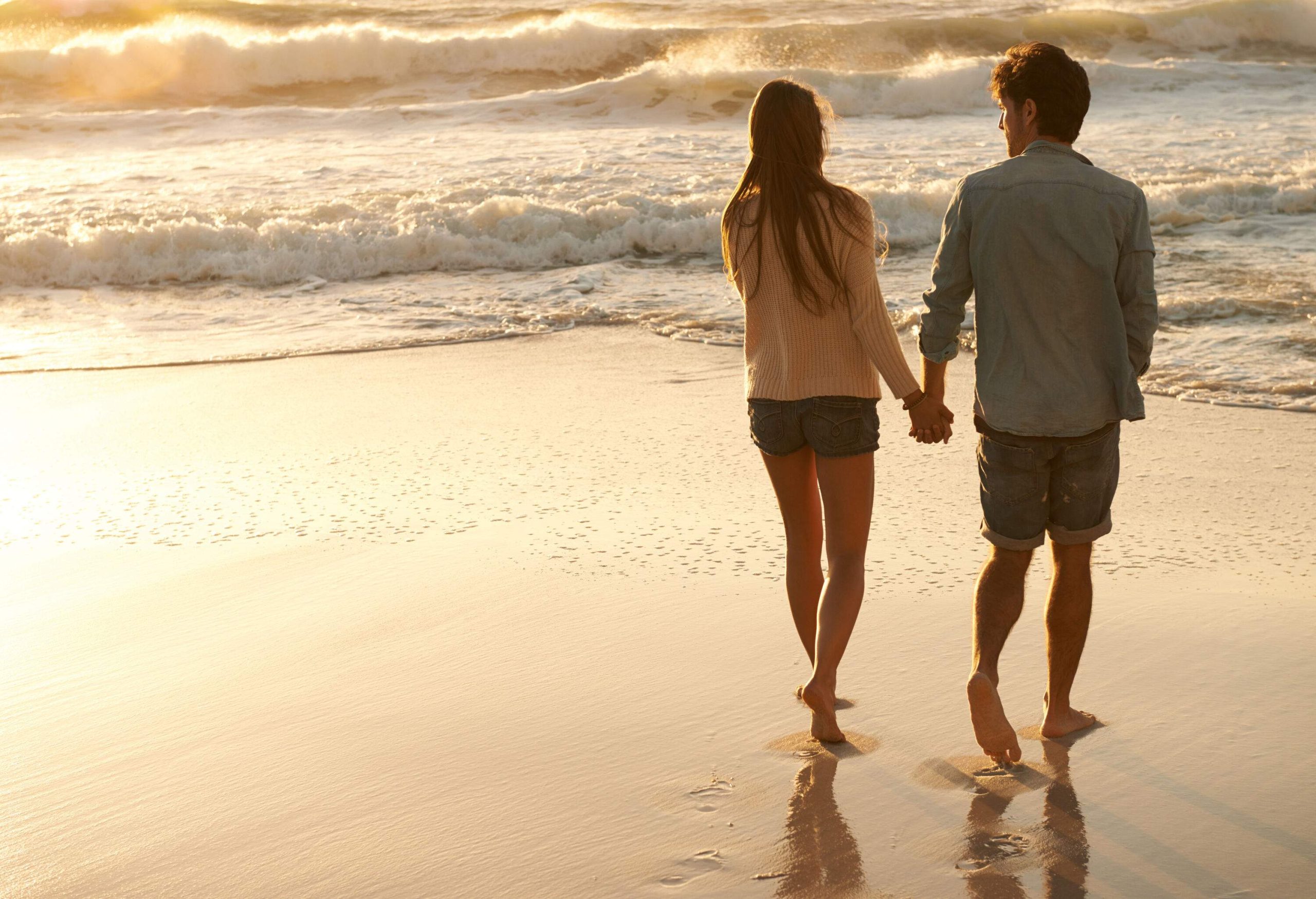 Back view of a couple holdings hands while walking on the beach with the big white waves.