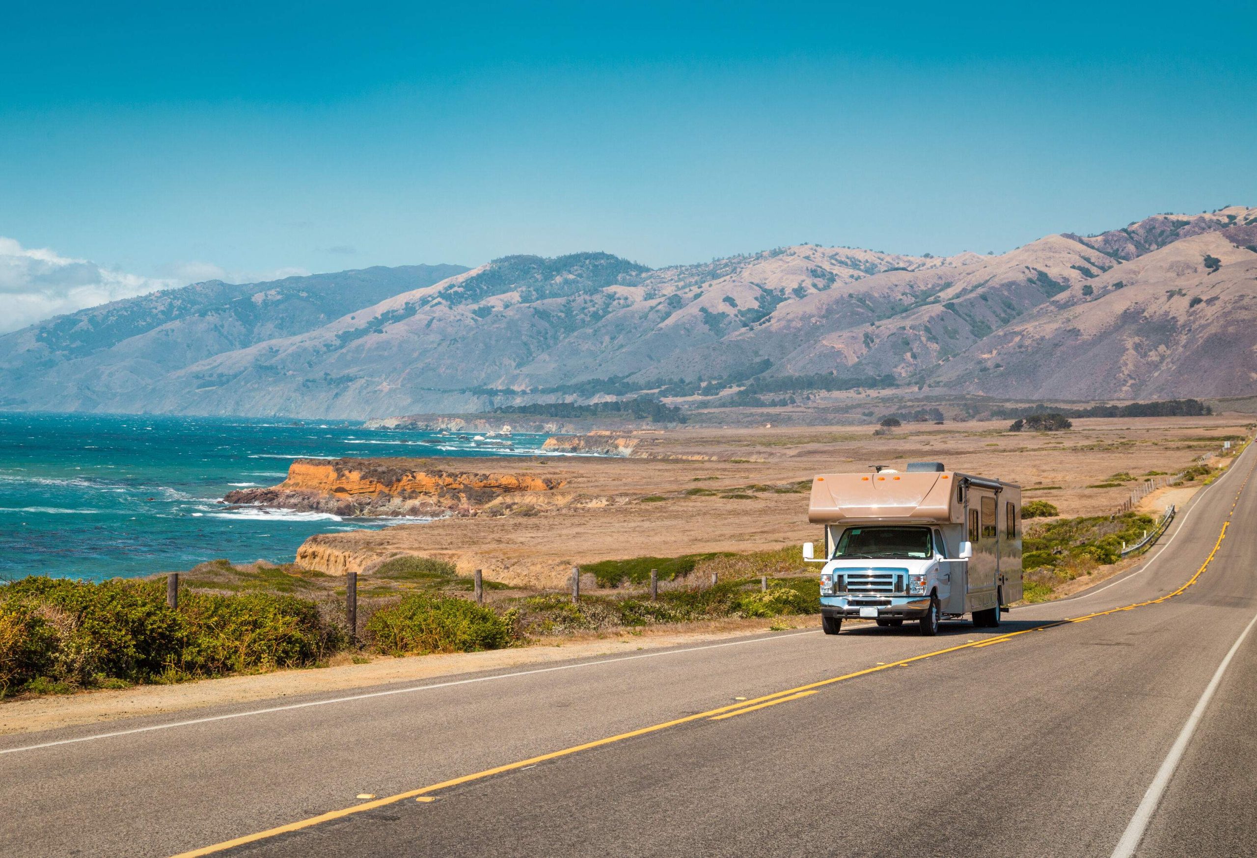 An RV moving along a road by the sea backed by a barren mountain range.