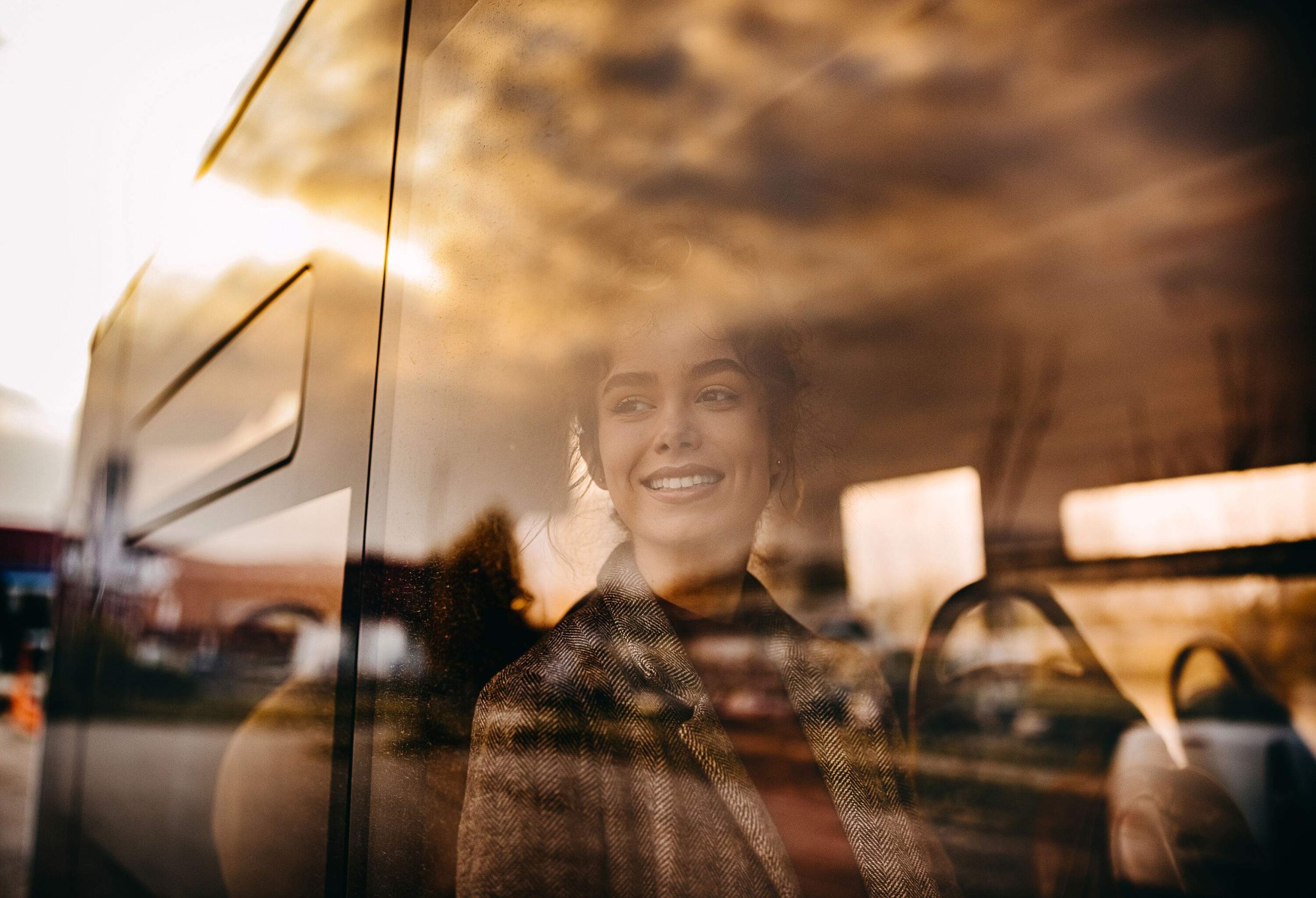 A woman in a coat smiles as she looks out a bus window.