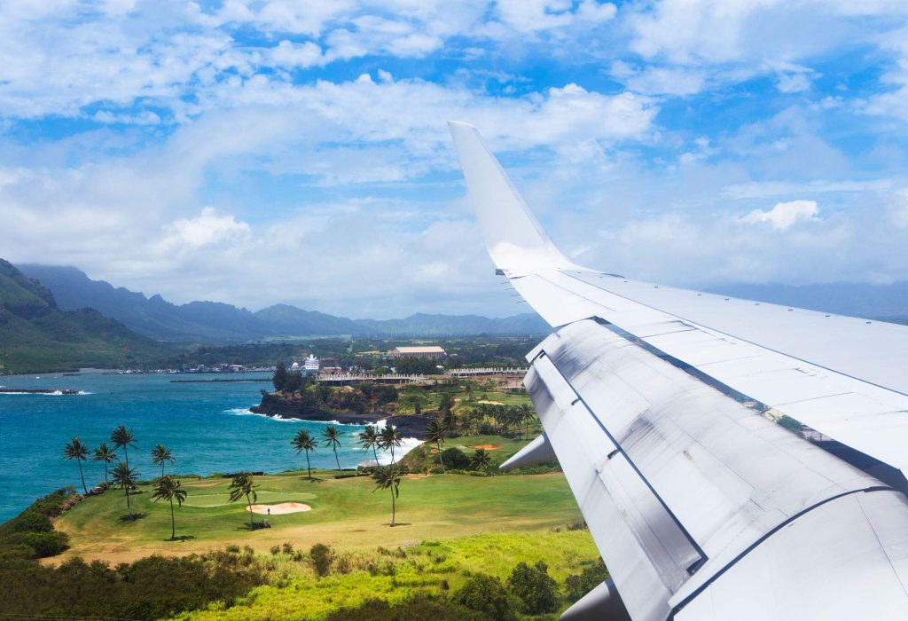 The expansive wing of a plane glides through the clear blue skies, with a lush tropical island sprawled below and a majestic mountain range visible in the distance.