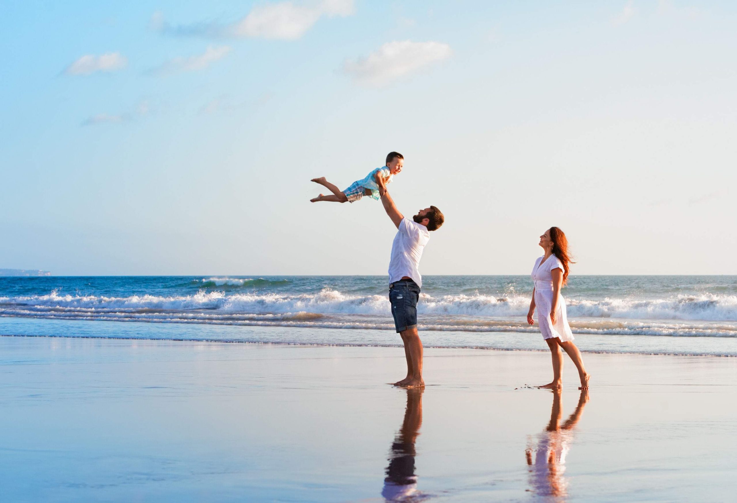 A man tossing a young boy in the air as a woman watches from behind them on the beach.