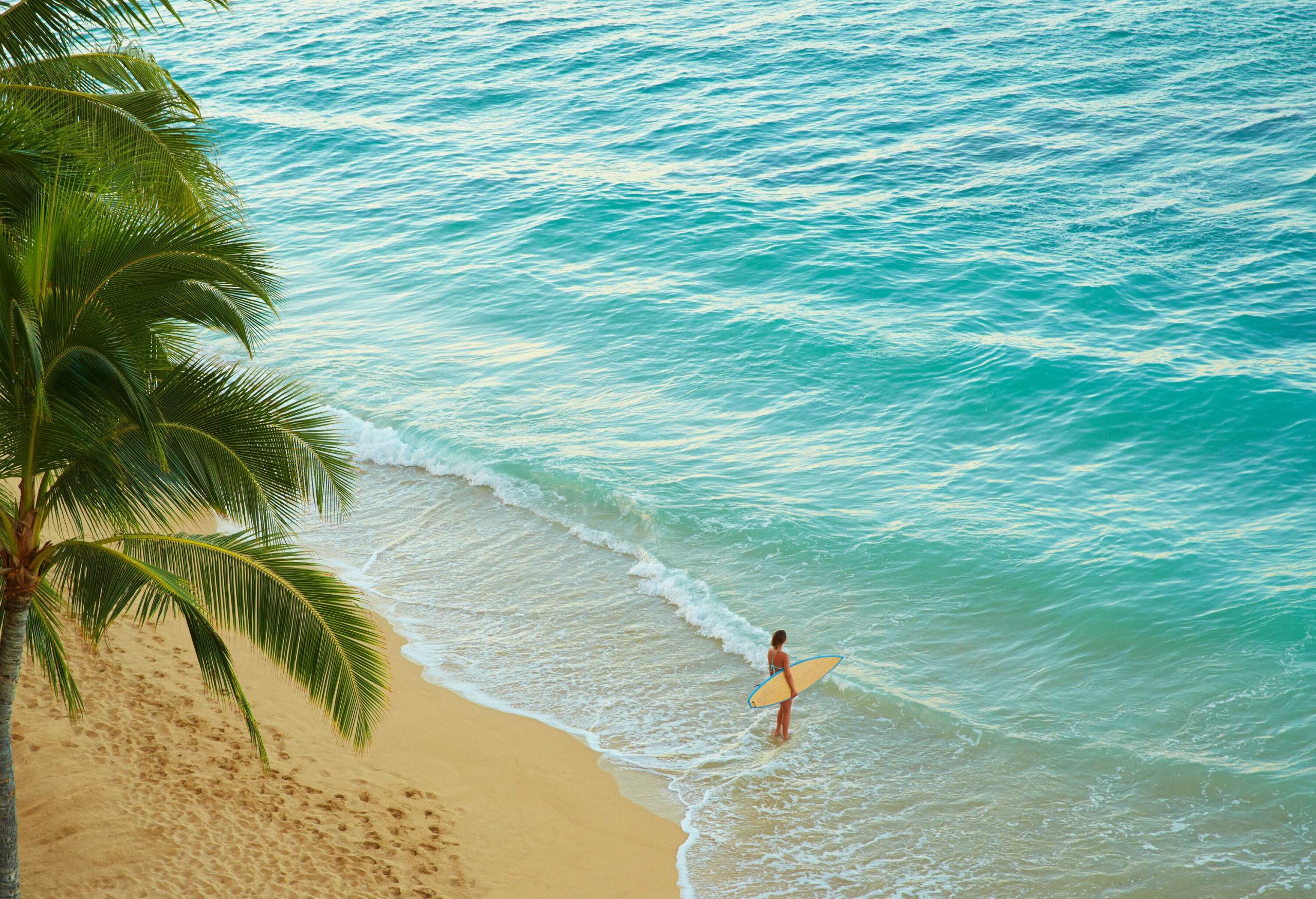 A woman standing on the beach with a surfboard while the waves smash on her feet.