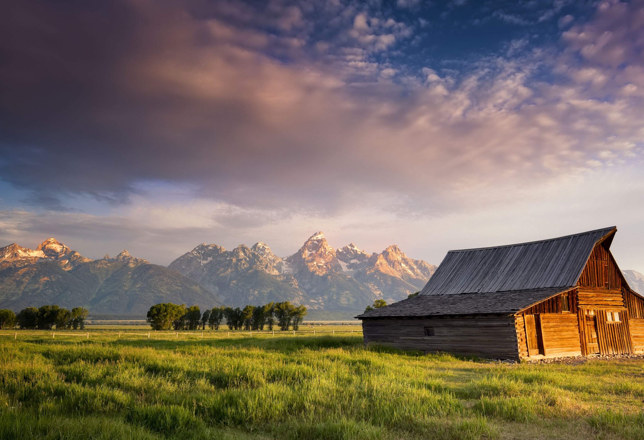 An old wooden barn in the lush ranchland bounded by a sharp-pointed, snow-capped mountain range.