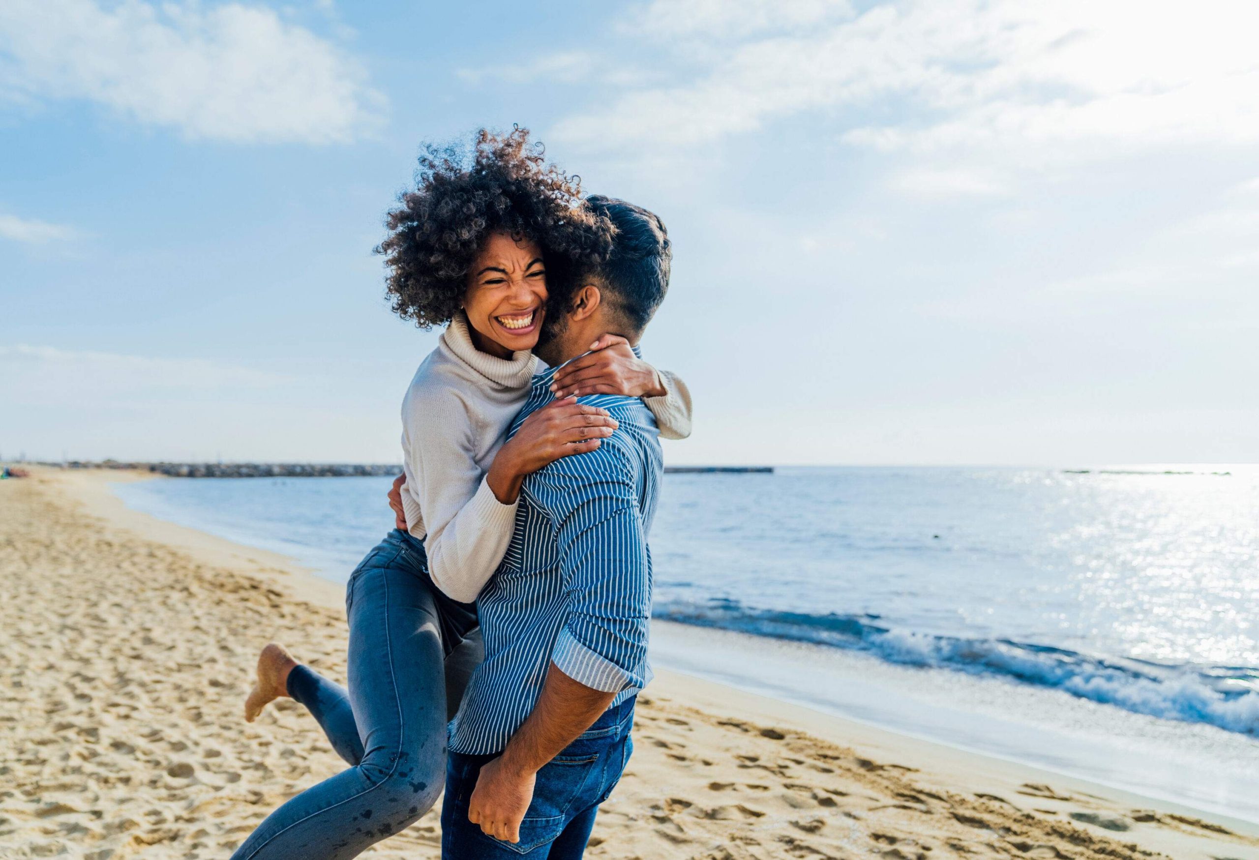 A curly-haired woman jumps and hugs a man on the beach.