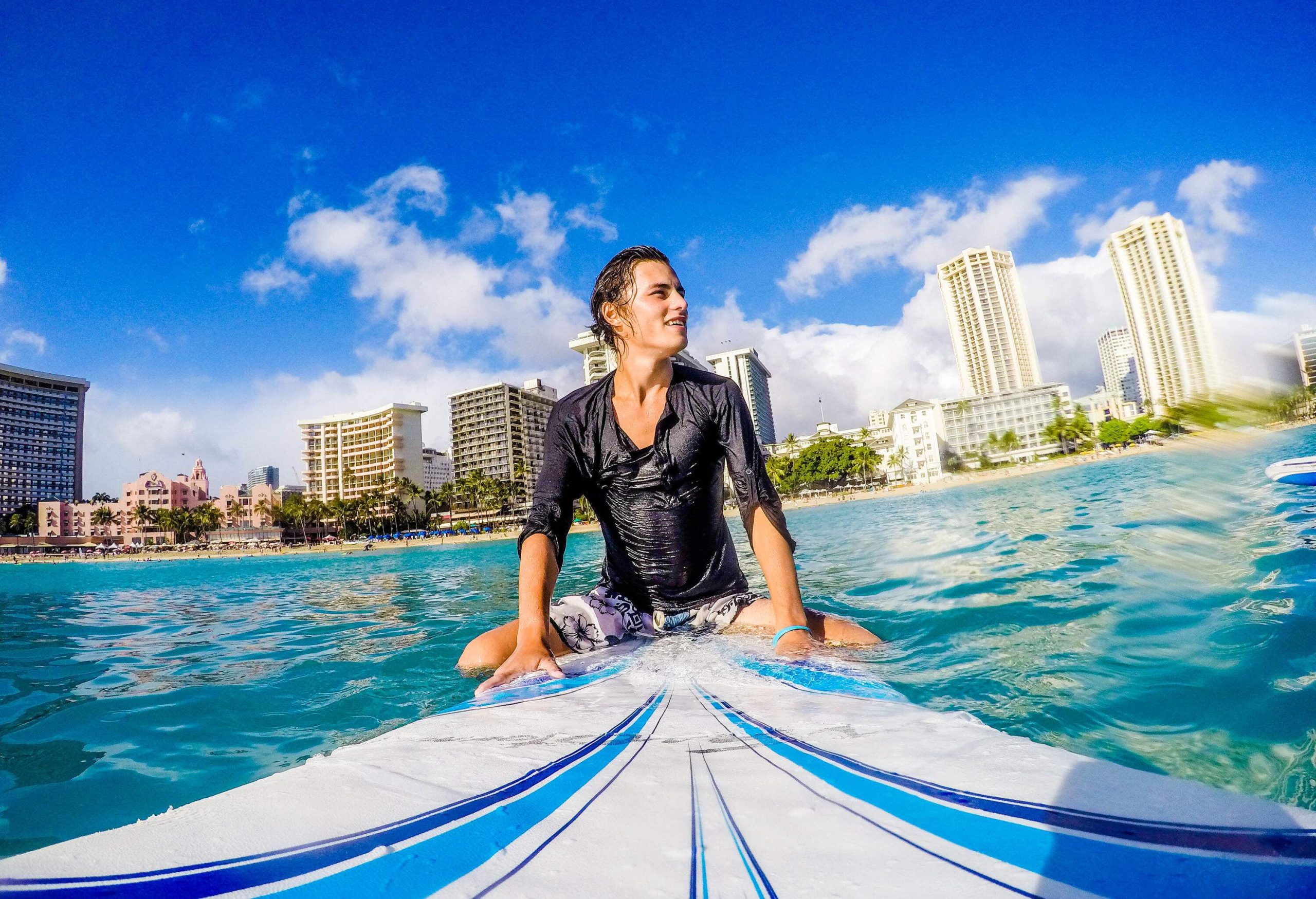 A person sits serenely on a surfboard atop the water, gazing into the distance, with a backdrop of buildings adding a touch of urban contrast to the peaceful coastal scene.