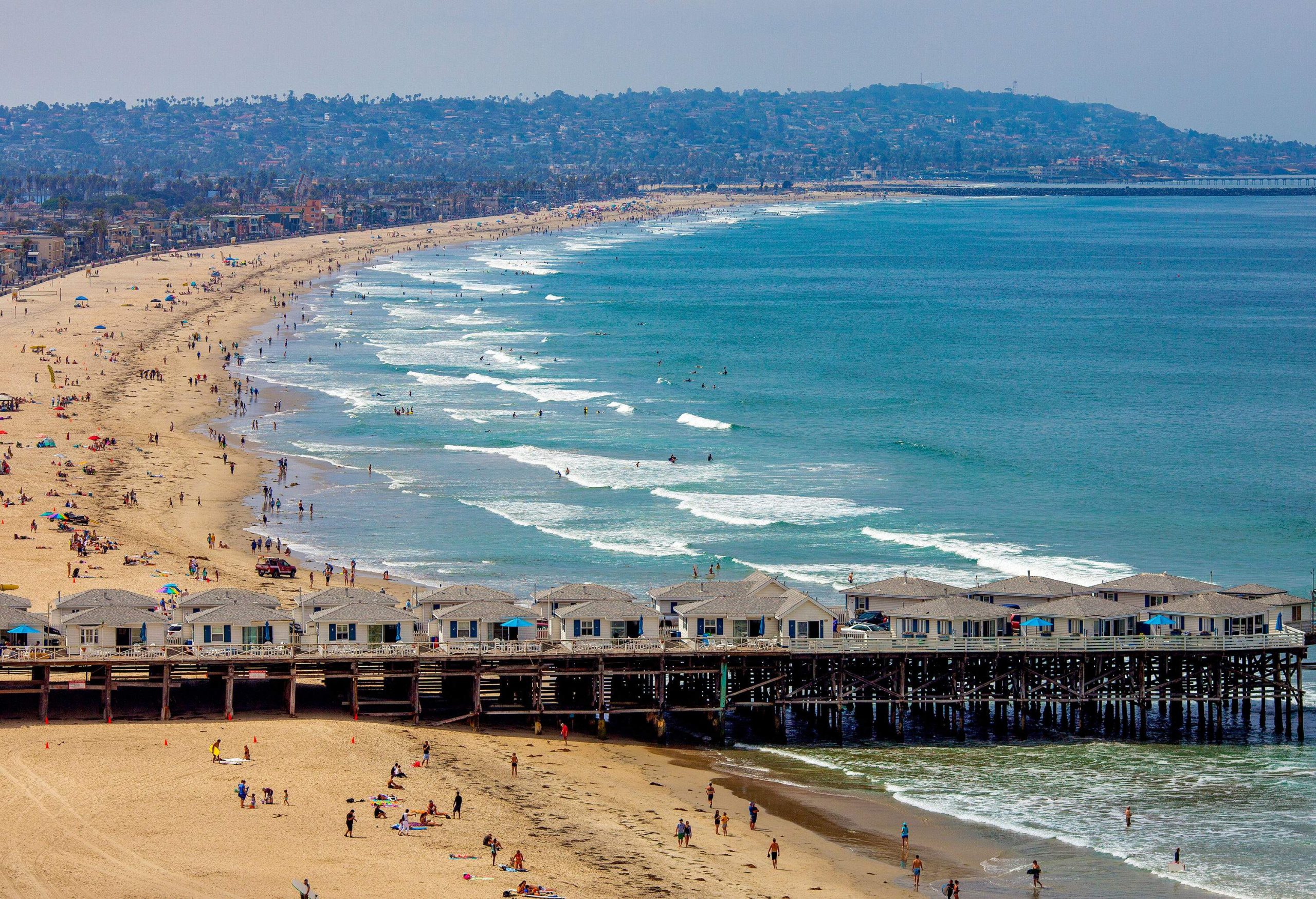 An aerial view of a bustling sandy beach with a pier adorned with hotel cottages.