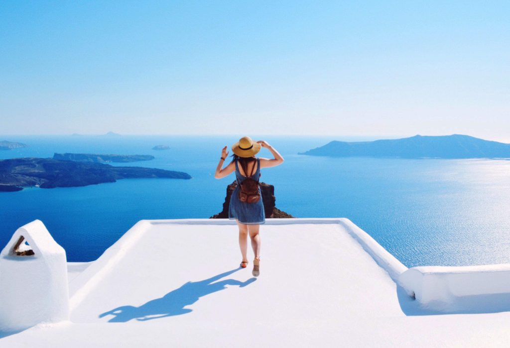 A woman in a blue dress stands on top of a whitewashed building overlooking a calm blue sea.