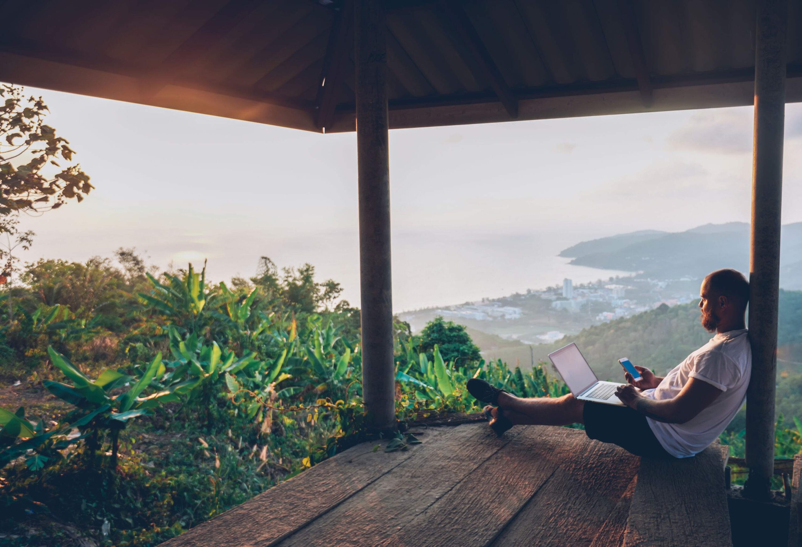 A male tourist sits on a wooden shed while working on his laptop and phone with a view of a village and the ocean downhill.