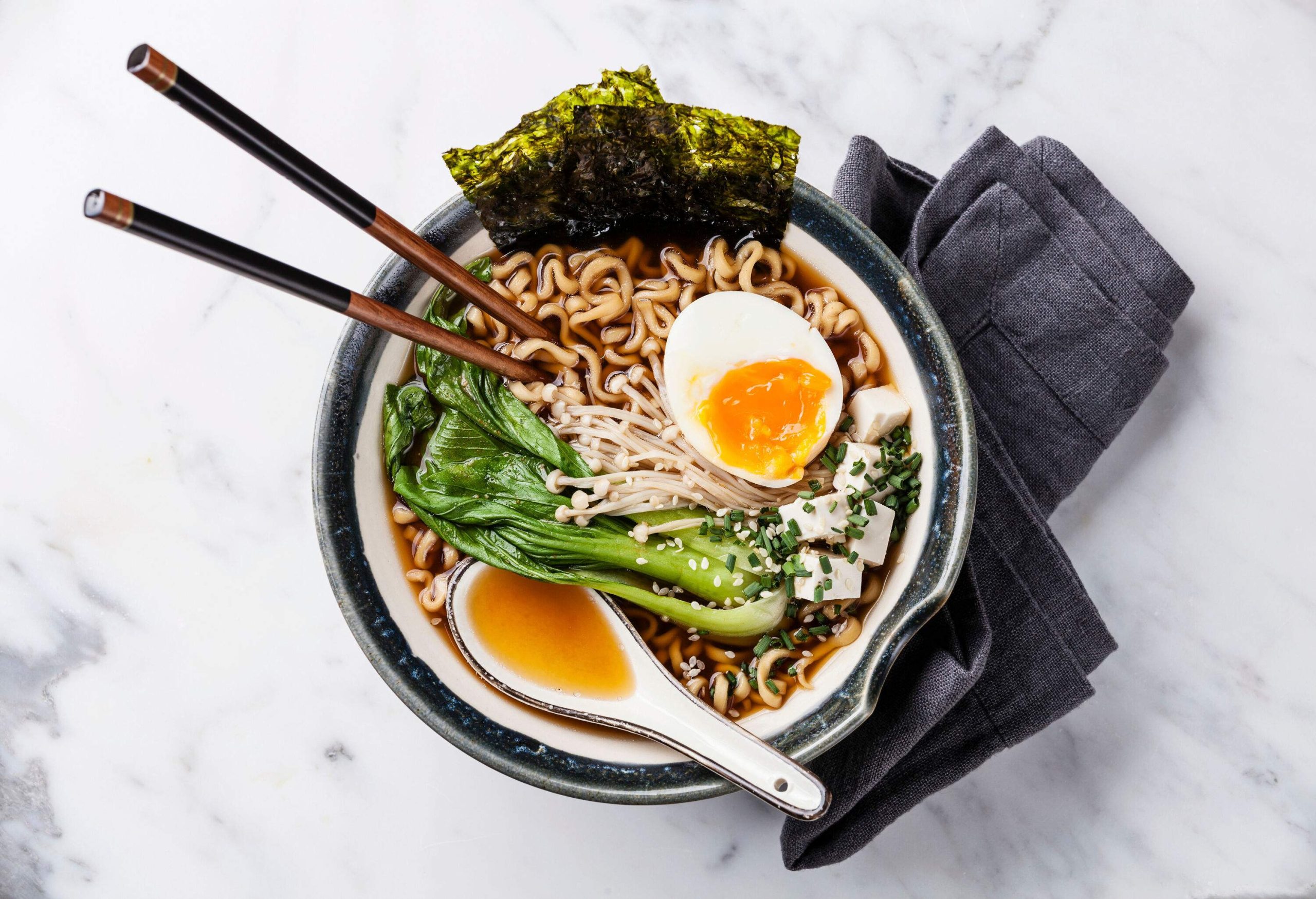 A bowl of ramen with seaweed sheets, soft-boiled egg, enoki mushroom, and bok choy on top of thick noodles.