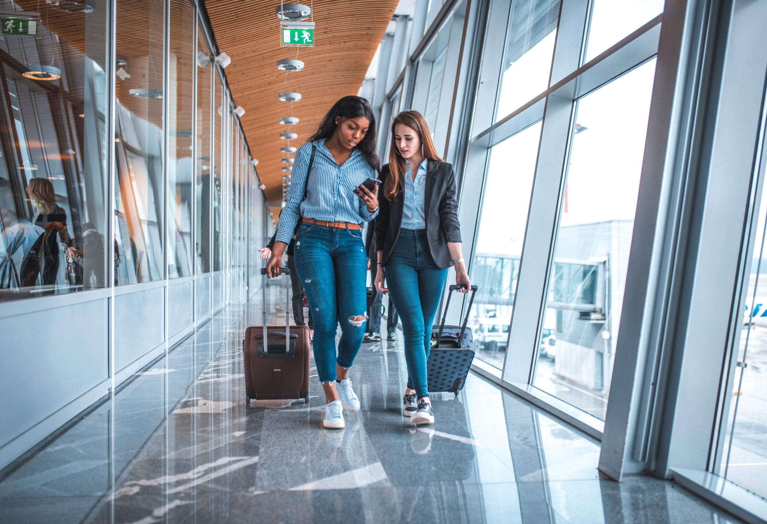 Two women dressed fashionably chic dragging their luggage through an airport terminal while checking on a smartphone.