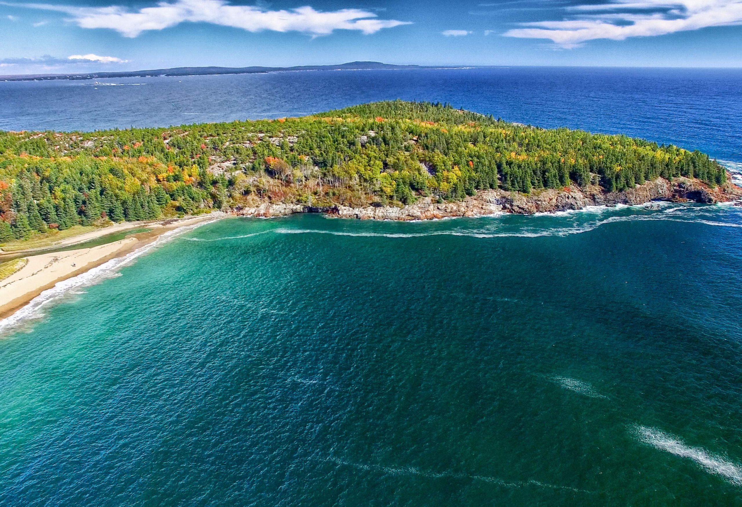 The deep blue sea smashing against the sand and the rocks bordering a forested island.