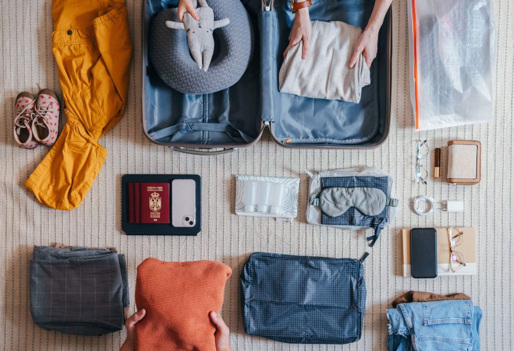 Flat lay of an anonymous family packing various things and essentials for their upcoming trip or vacation. Unrecognizable little girl putting in a toy in the suitcase. There is passport, mobile phone, travel pillow and other things laid on the carpet on the floor.