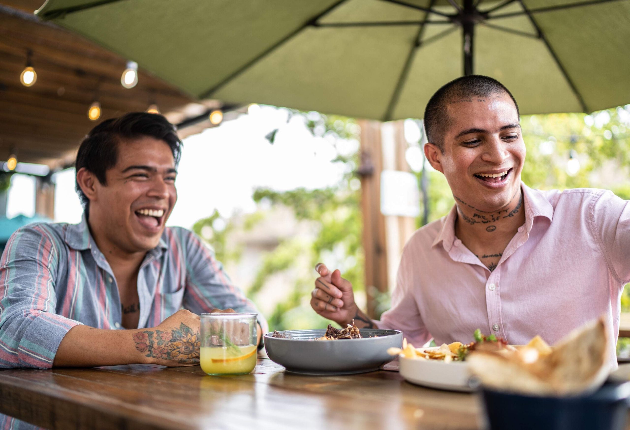 Male lovers laughing at something while sitting at a table with food and beverages.