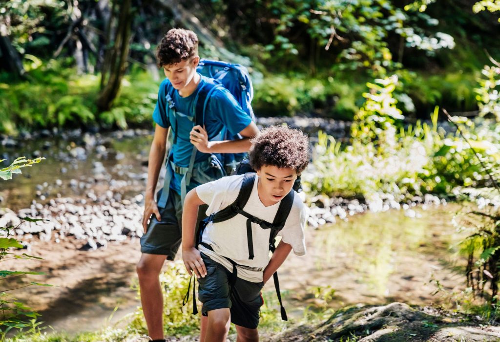 Two boys with backpacks traverse a river trail in the forest.