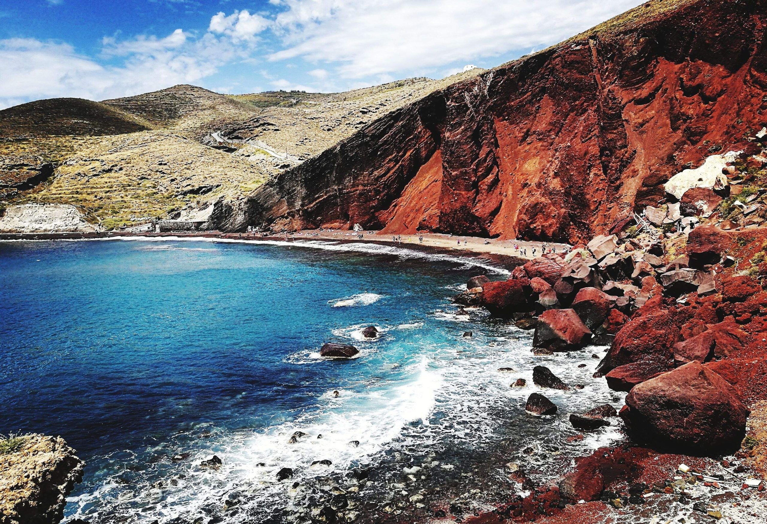 A beach with big boulders along a red steep cliff.