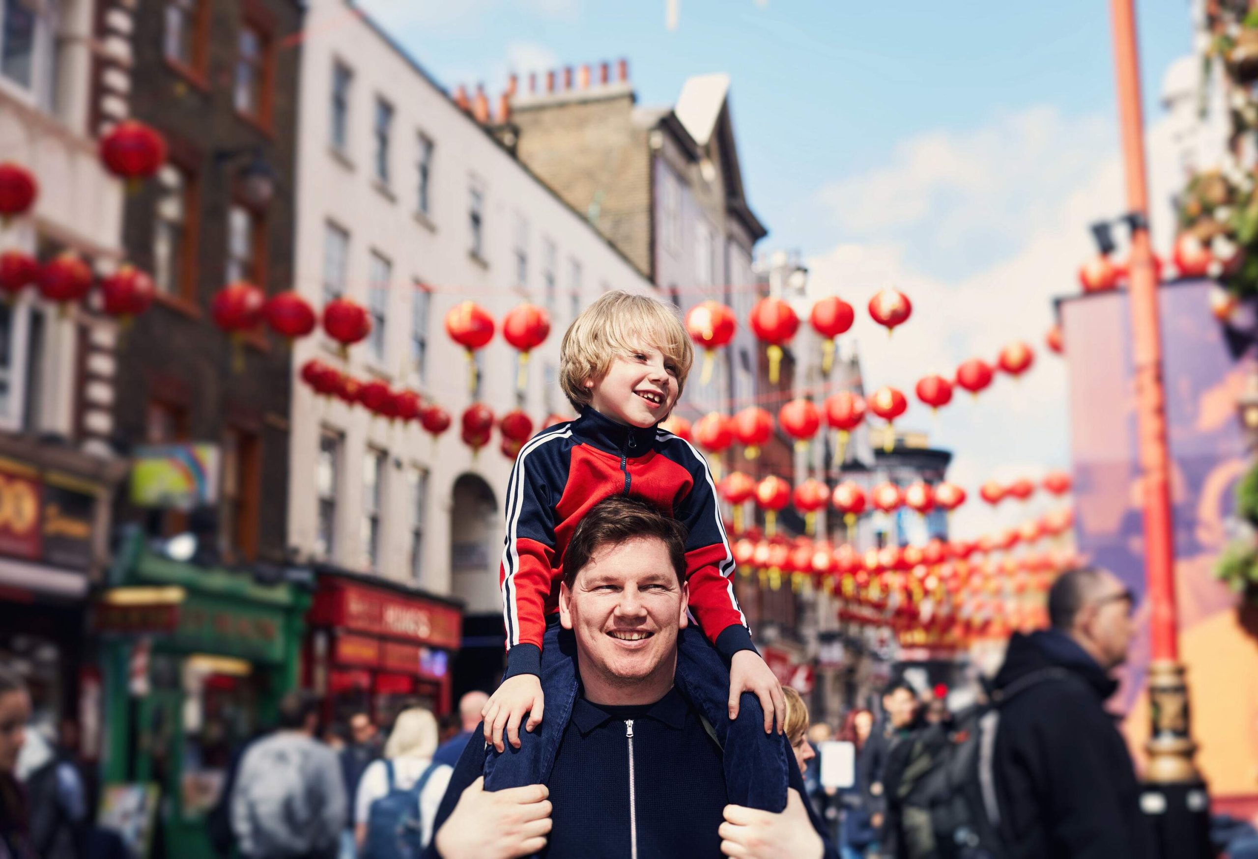 A man with his son on his shoulders walking down the street with red lanterns hanging overhead.