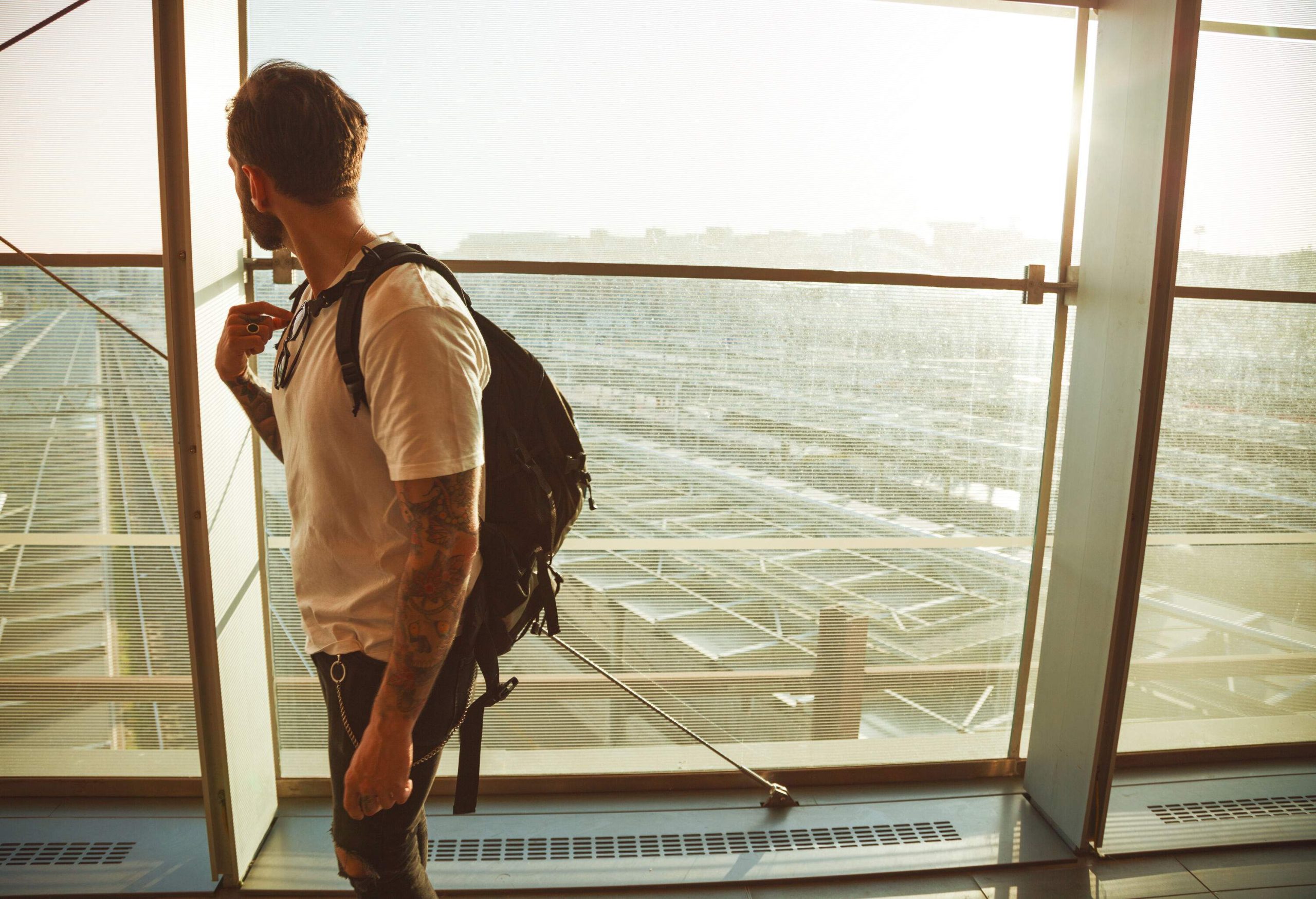 A tattooed man with a backpack looking out the large glass windows of an airport terminal.